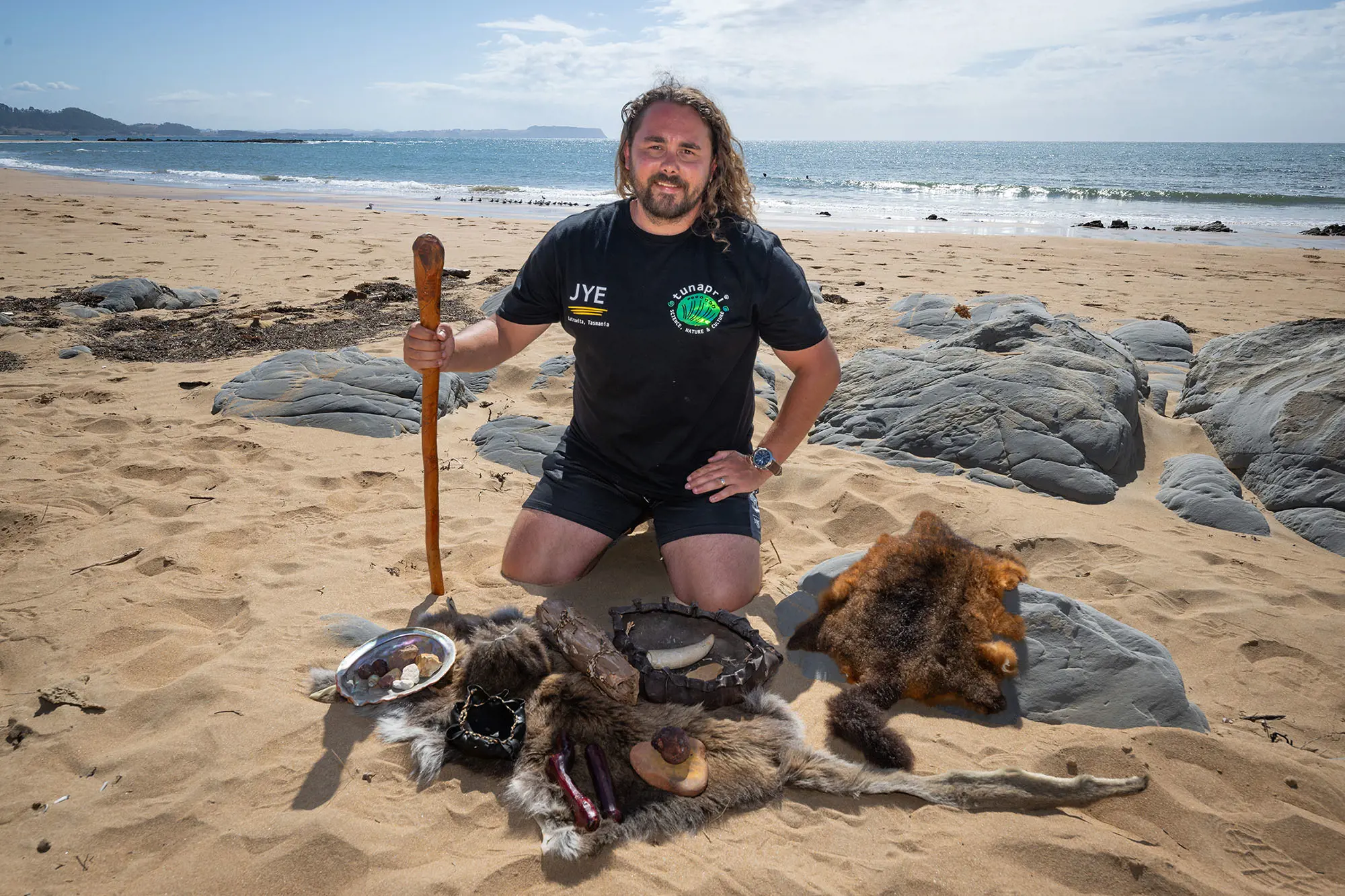 A man kneels on a sandy beach, with a spread of Tasmanian Aboriginal items like kelp baskets and shells lying on a wallaby skin in front of him. His black shirt has a business logo on one side and 'JYE' on the other.