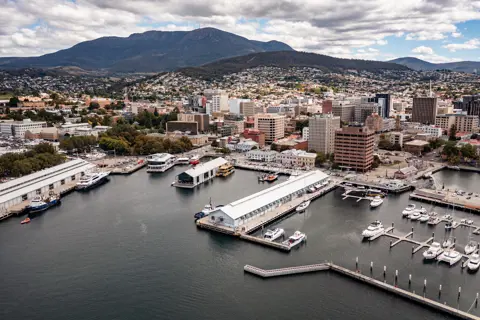 An aerial view of Hobart's waterfront, with ships moored, long piers, the CBD buildings and beyond, a mountainous landscape.