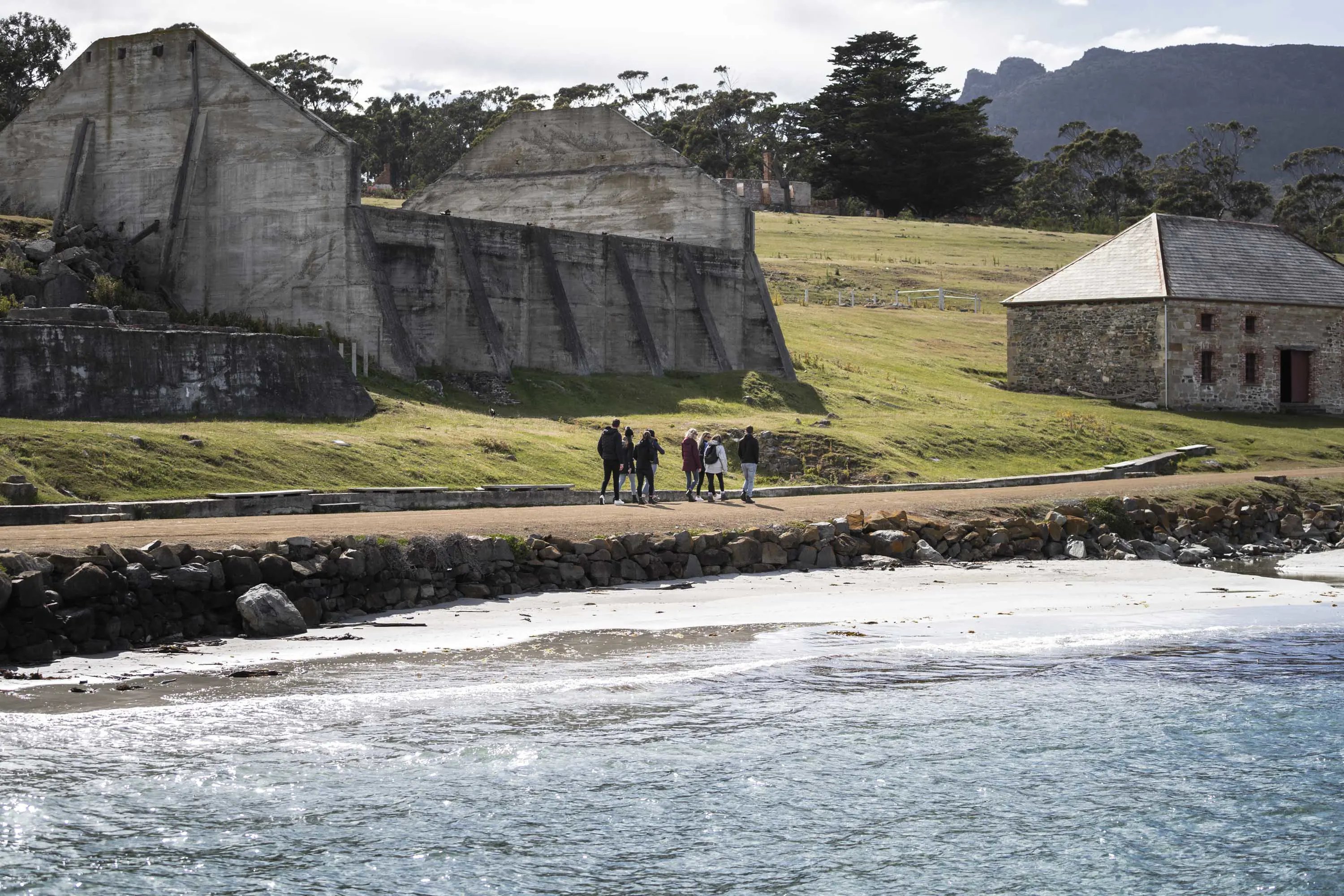 Large concrete walls of a factory ruin stand above a gravel path around a bay.