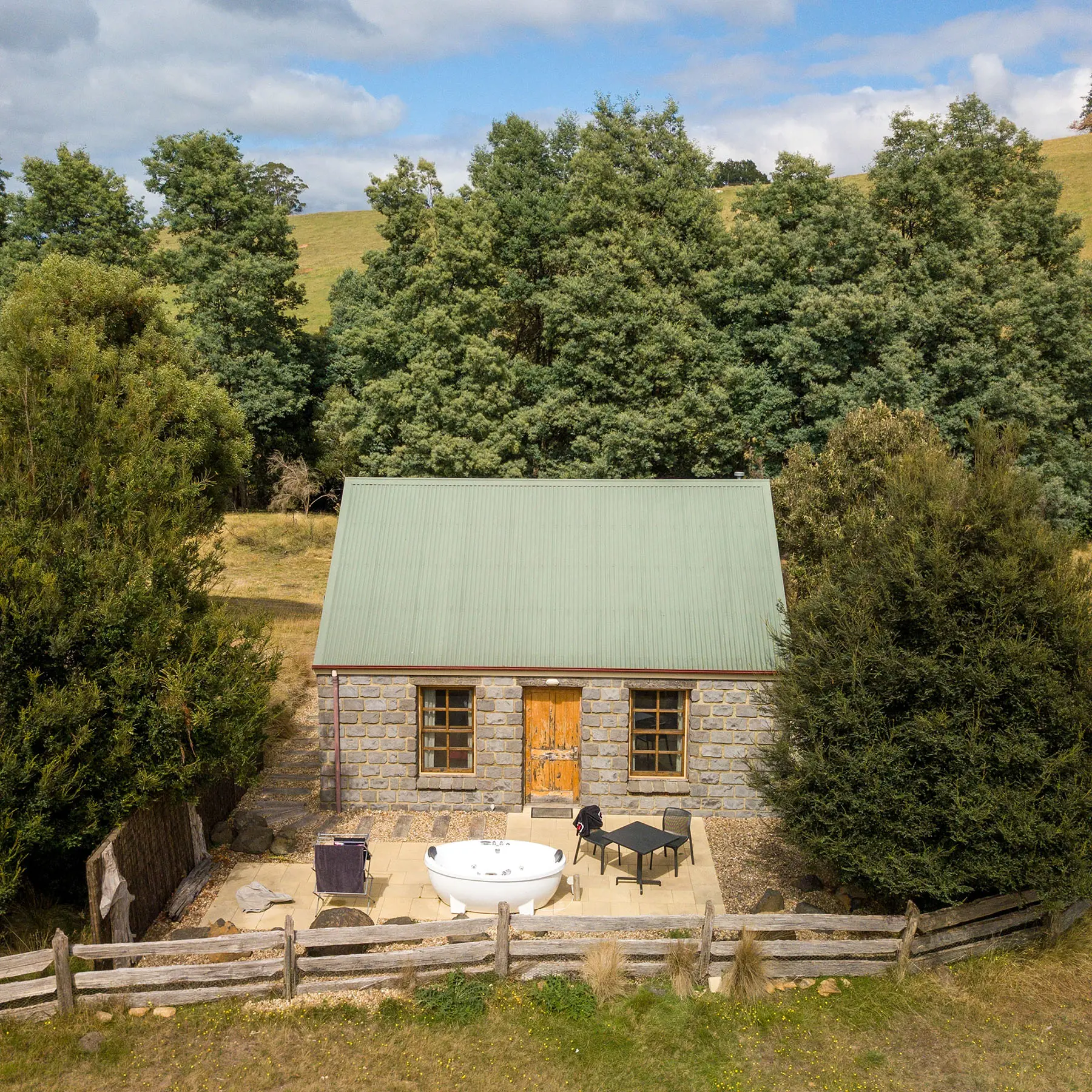 A small rustic brick cottage nestled into the trees. Out the front there is a spacious white outdoor bath and some patio furniture.
