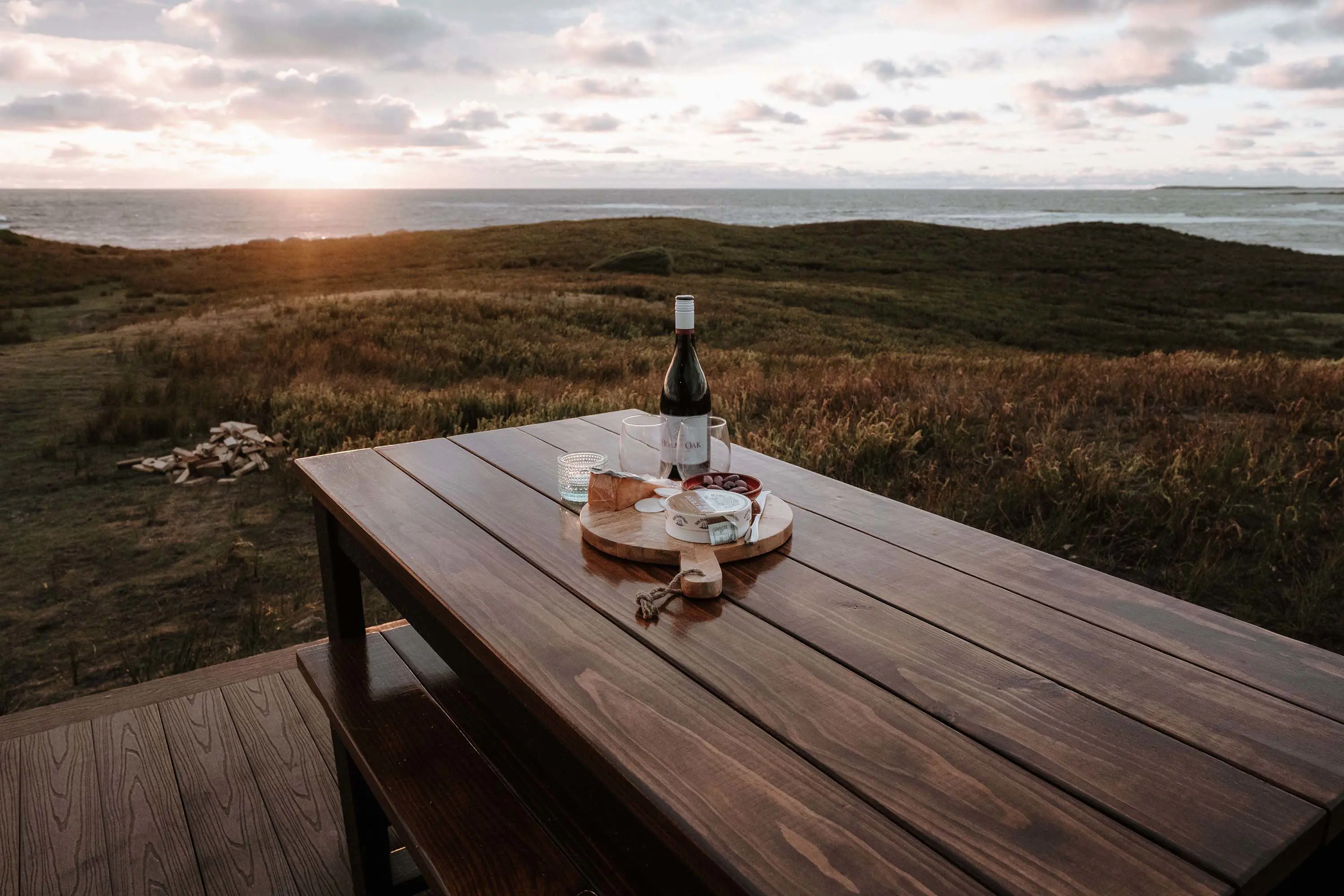 A long wooden table stands on an outdoor deck, looking over rolling grass hills to the ocean beyond. On the table is a cheese and wine platter.