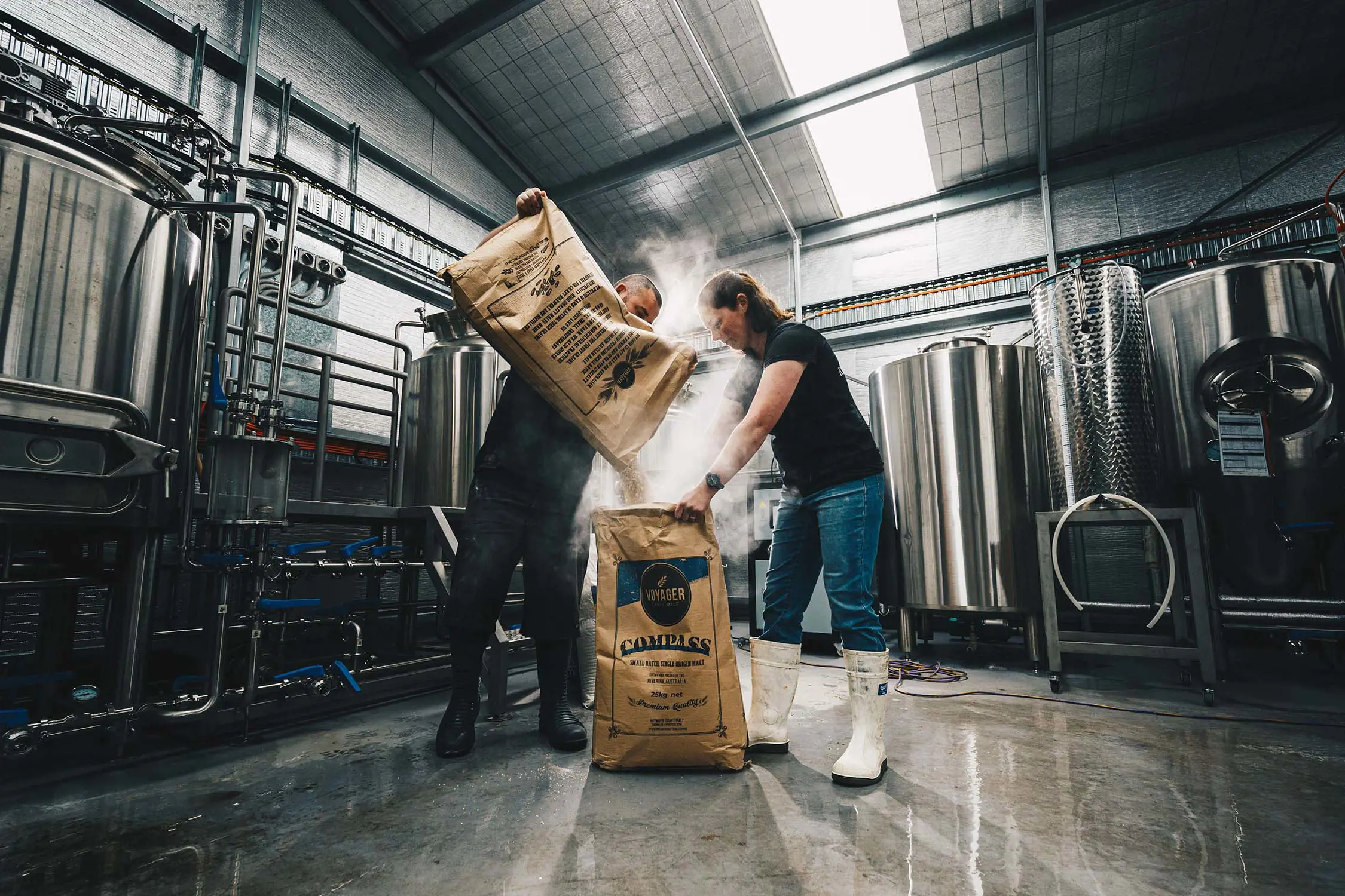 Two people stand in a large industrial kitchen-like space, surrounded by large silver stills and pipes. The woman holds a brown paper bag of flour open on the ground and the man is pouring the contents of another bag into it, sending a cloud of flour into the air.