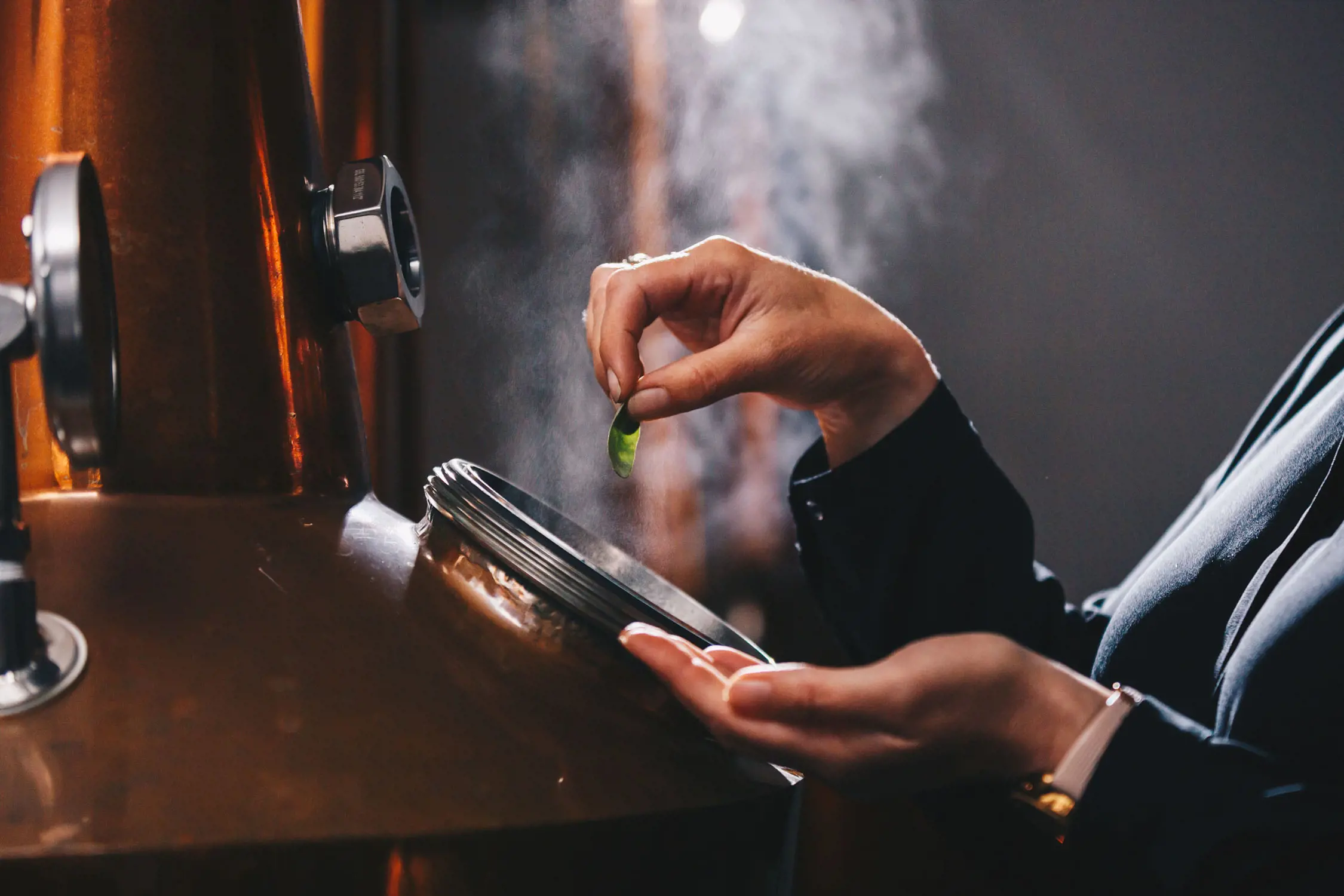 A person's hand drops a leaf into an open valve of a copper distilling still, with steam rising from the opening.