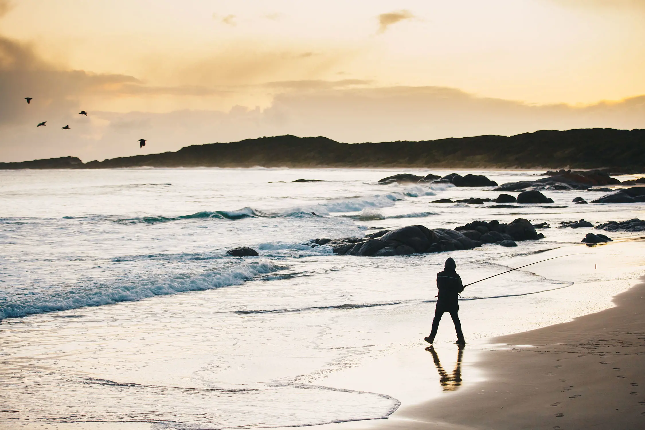 At a rugged beach covered with rocks, rolling waves and overlooked by dramatic hills, a person casting a fishing line is silhouetted on the sand.