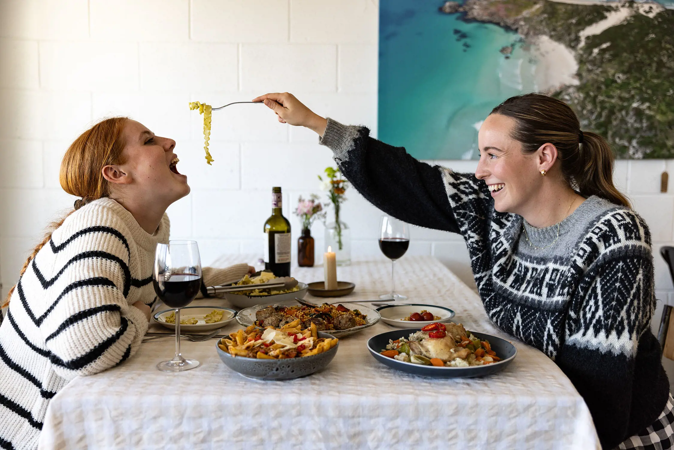 Two women sit either side of a table covered with a white tablecloth and laden with plates of delicious food and red wine. One holds out a fork with strands of pasta and the other leans forward with her mouth open, ready to try.