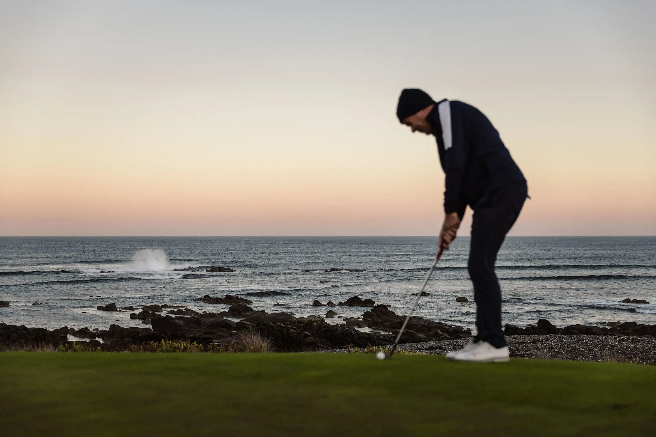 A man stands on a golf green, overlooking a dramatic ocean vista, with waves crashing onto the rocks. He is bent slightly, holding a golf club up to a ball on the grass, ready to swing.