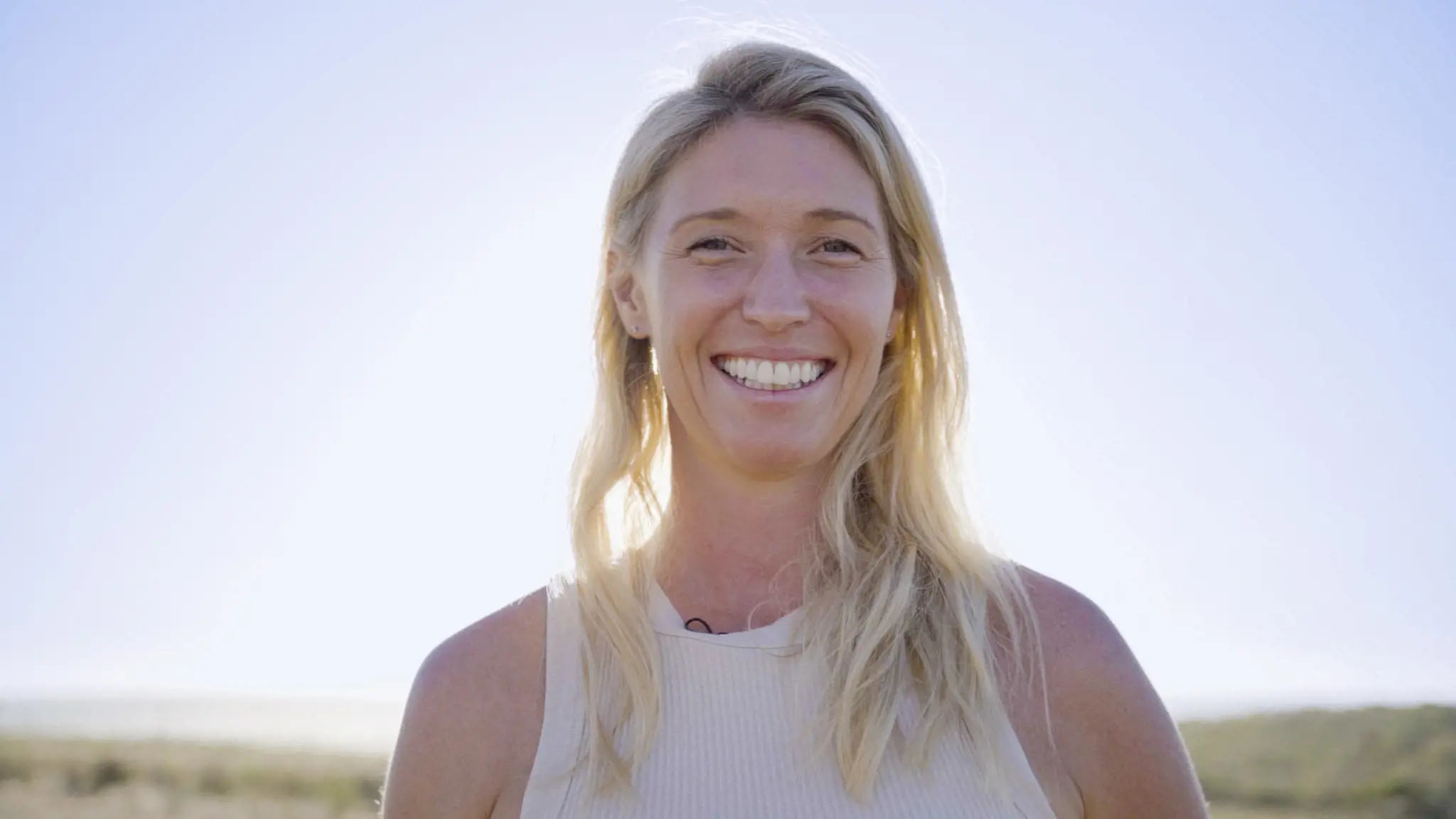 A woman with beach blonde hair and a white tank top stands on a coastal landscape, smiling for the camera.