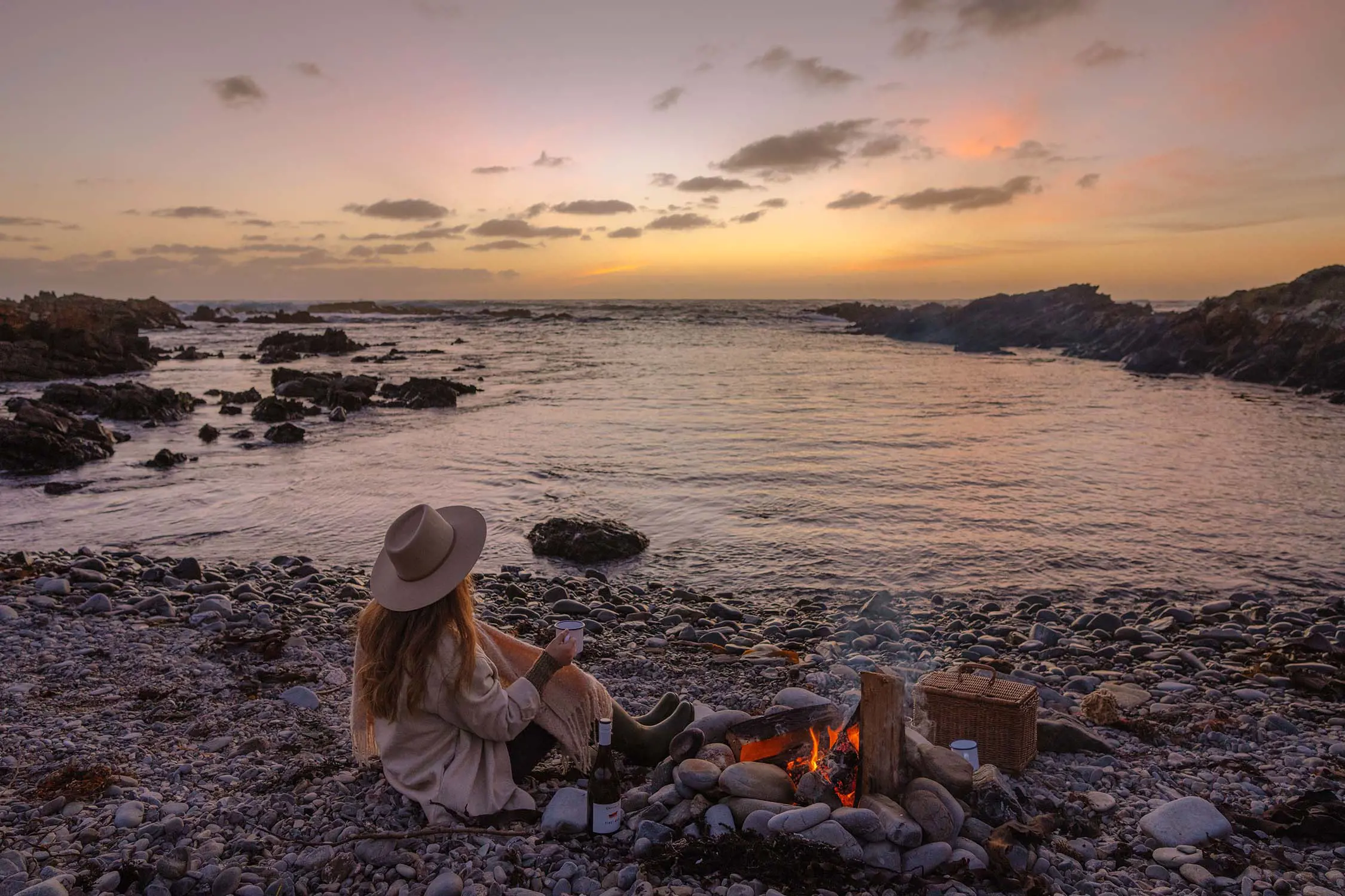 A woman sits on a rocky beach holding a hot drink, next to a small campfire. The sunset casts an orange-yellow glow over the scattered clouds in the sky.