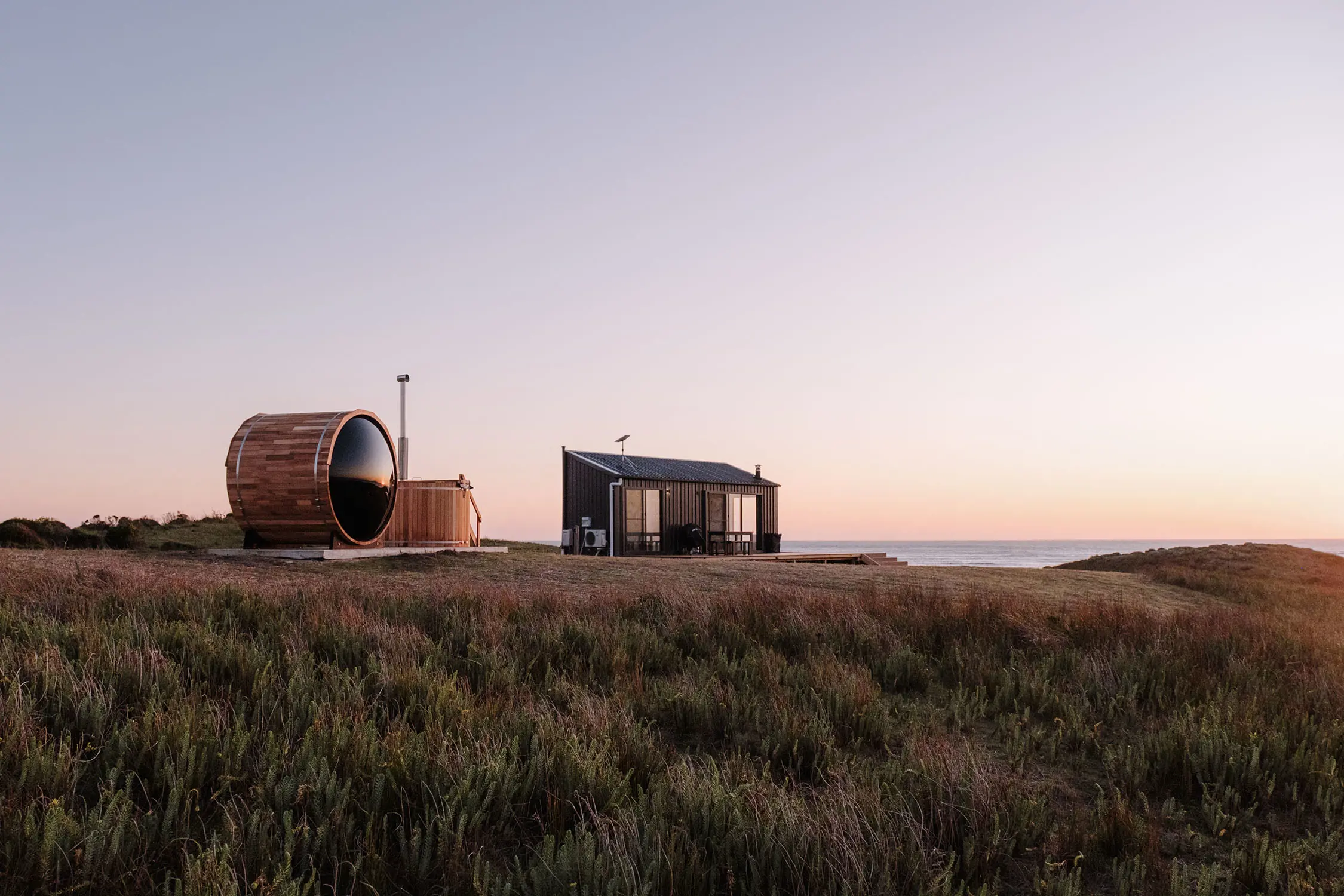 A modern, rustic cabin set in an open coastal field. Next to it is a large, barrel-shaped wooden sauna with a curved roof. The cabin and sauna face the ocean and the landscape is dotted with tall grasses swaying in the wind.