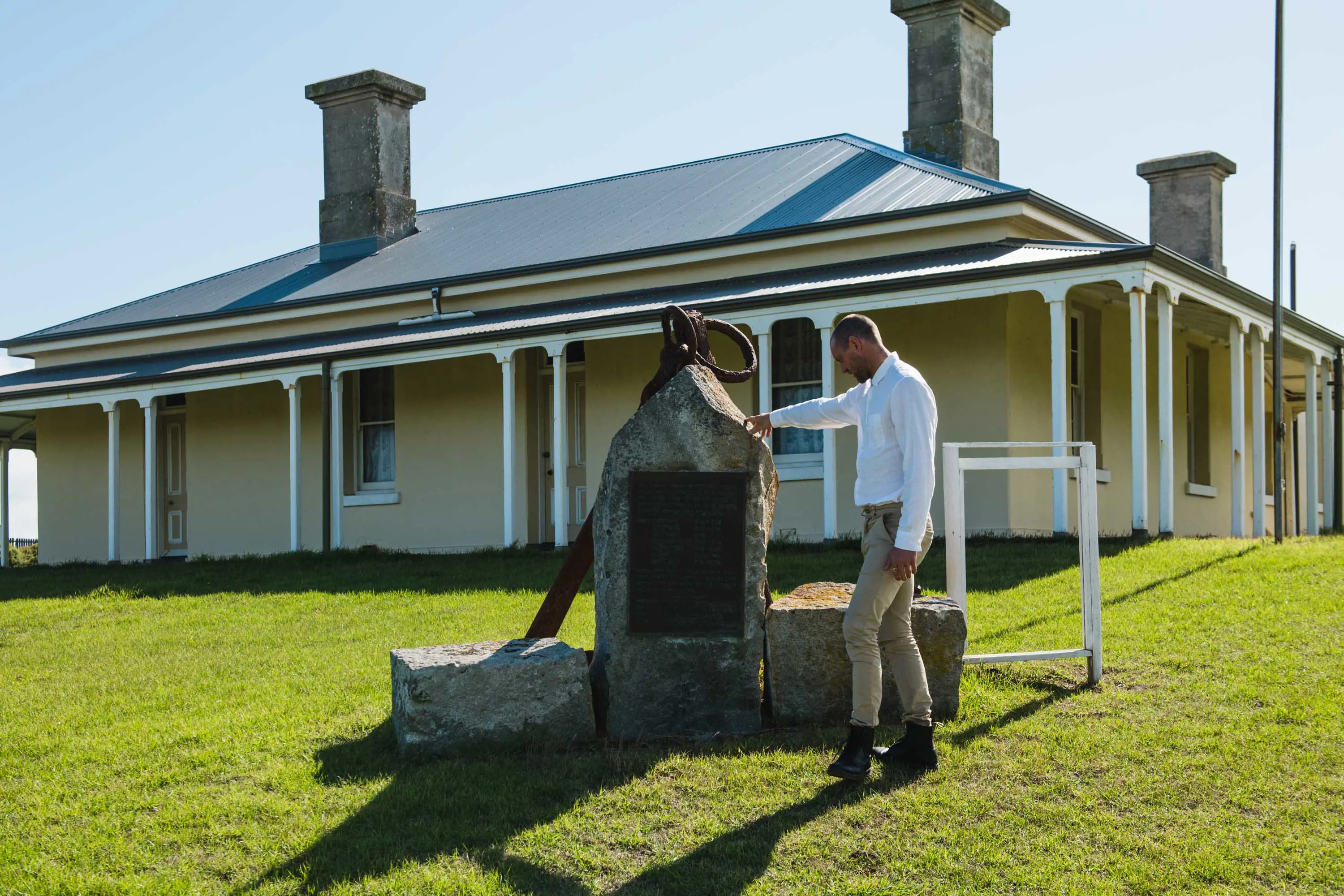A man wearing a white shirt, khaki pants and hiking boots stands next to a stone memorial in front of a colonial style house.