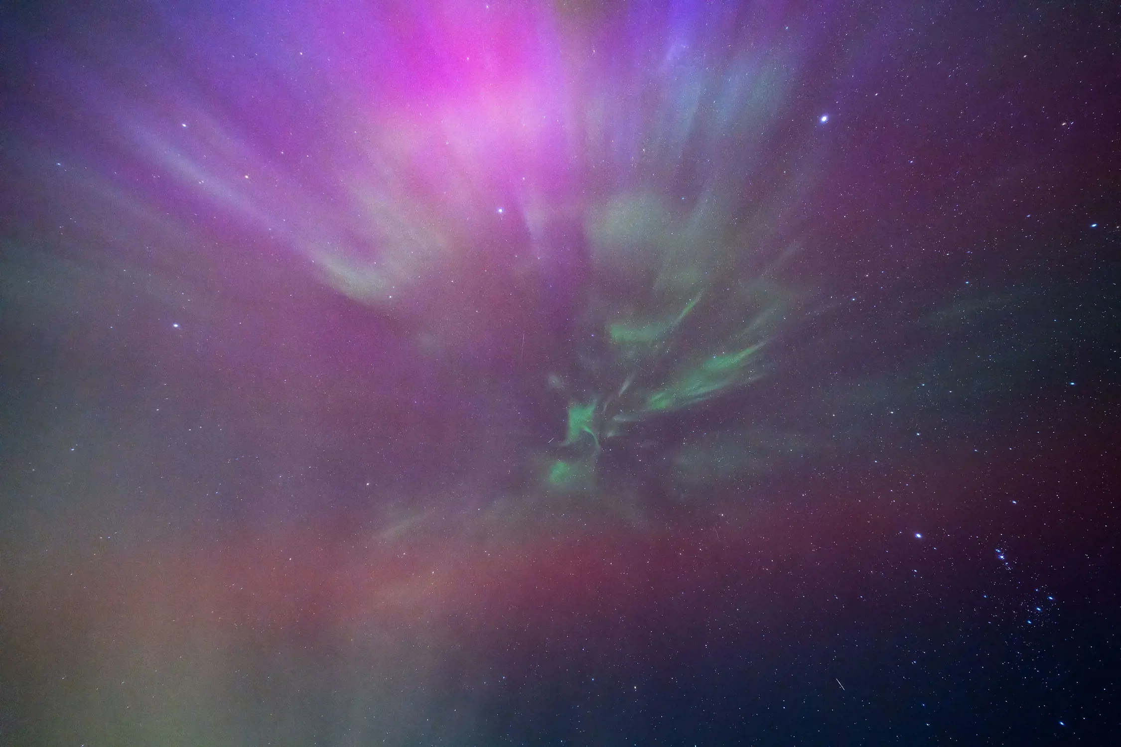 Looking up into the night sky, a brilliant, starburst-like view of aurora beams that glow green, purple and pink. Bright specks of stars shine through behind the beams.
