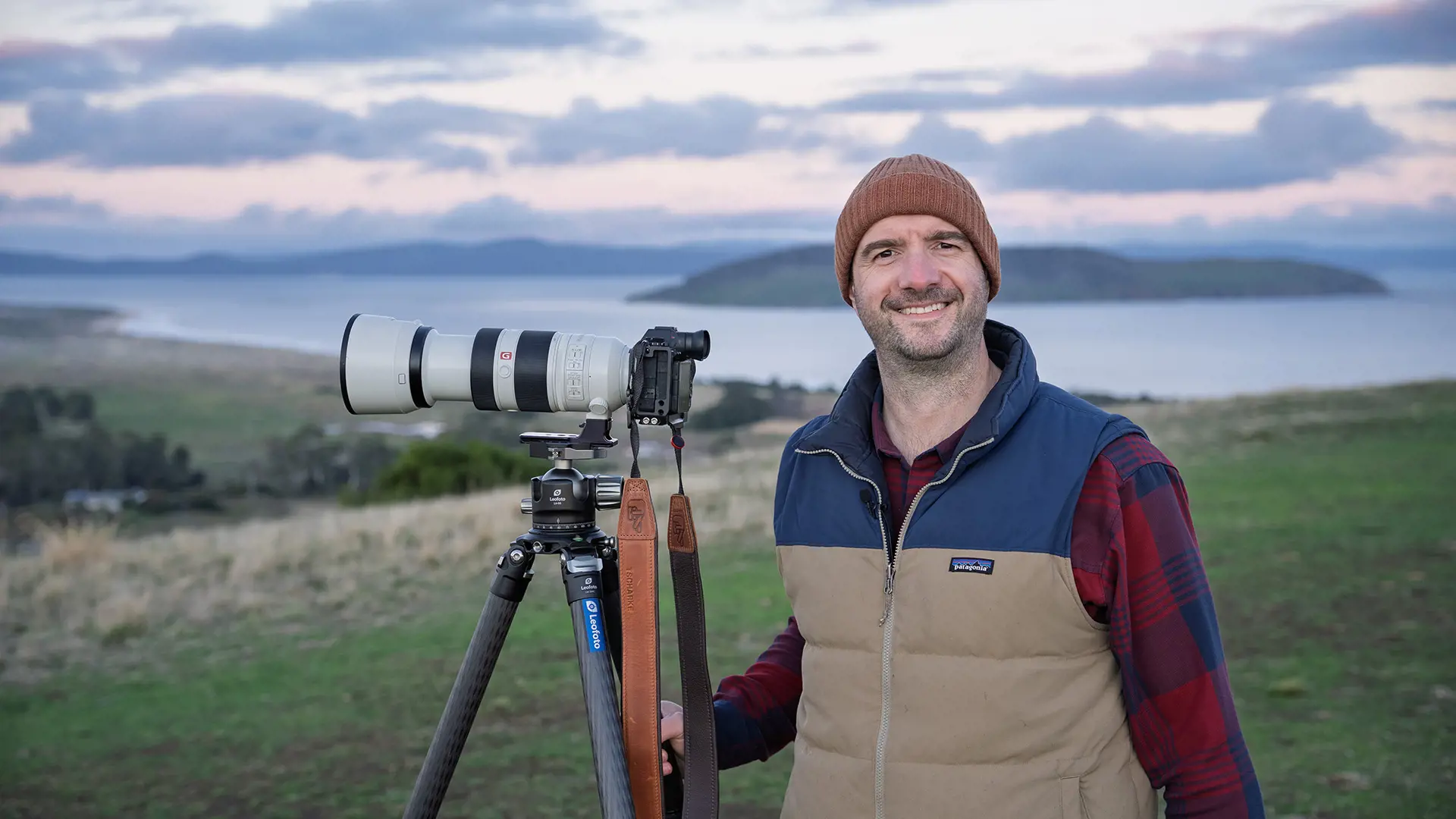 A man in a beanie and puffer vest stands on a hillside smiling for the camera. Next to him is a tripod with a large camera and lens attached.