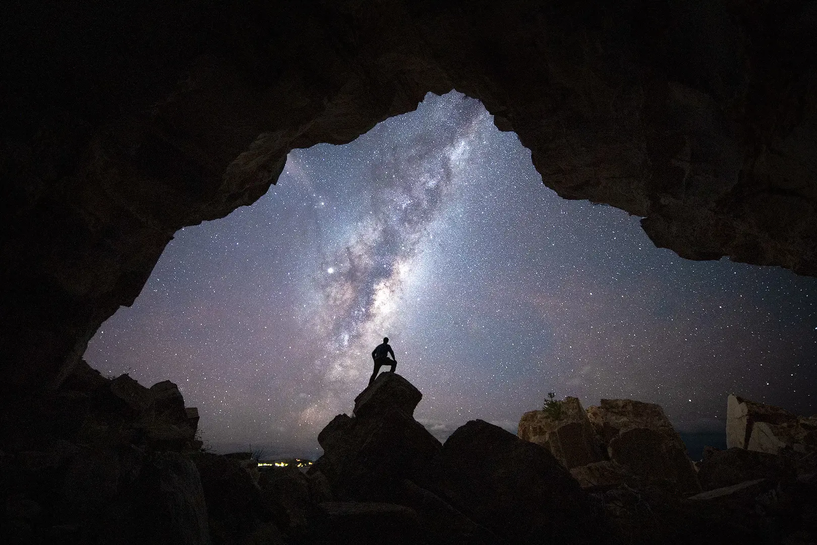 A stunning image of the night sky streaked through with the Milky Way, framed by the silhouette of the mouth of a cave. A person's silhouette is standing atop a rock, framed perfectly by the rocky surroundings.
