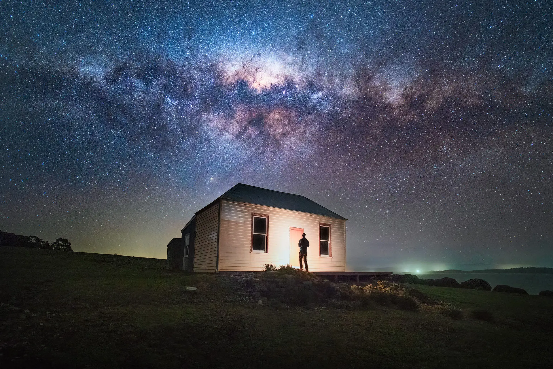 A vivid image of the Milky Way snaking across the night sky, over the top of an illuminated weatherboard cottage standing alone on a hill, with the silhouette of a person standing in front of the door.