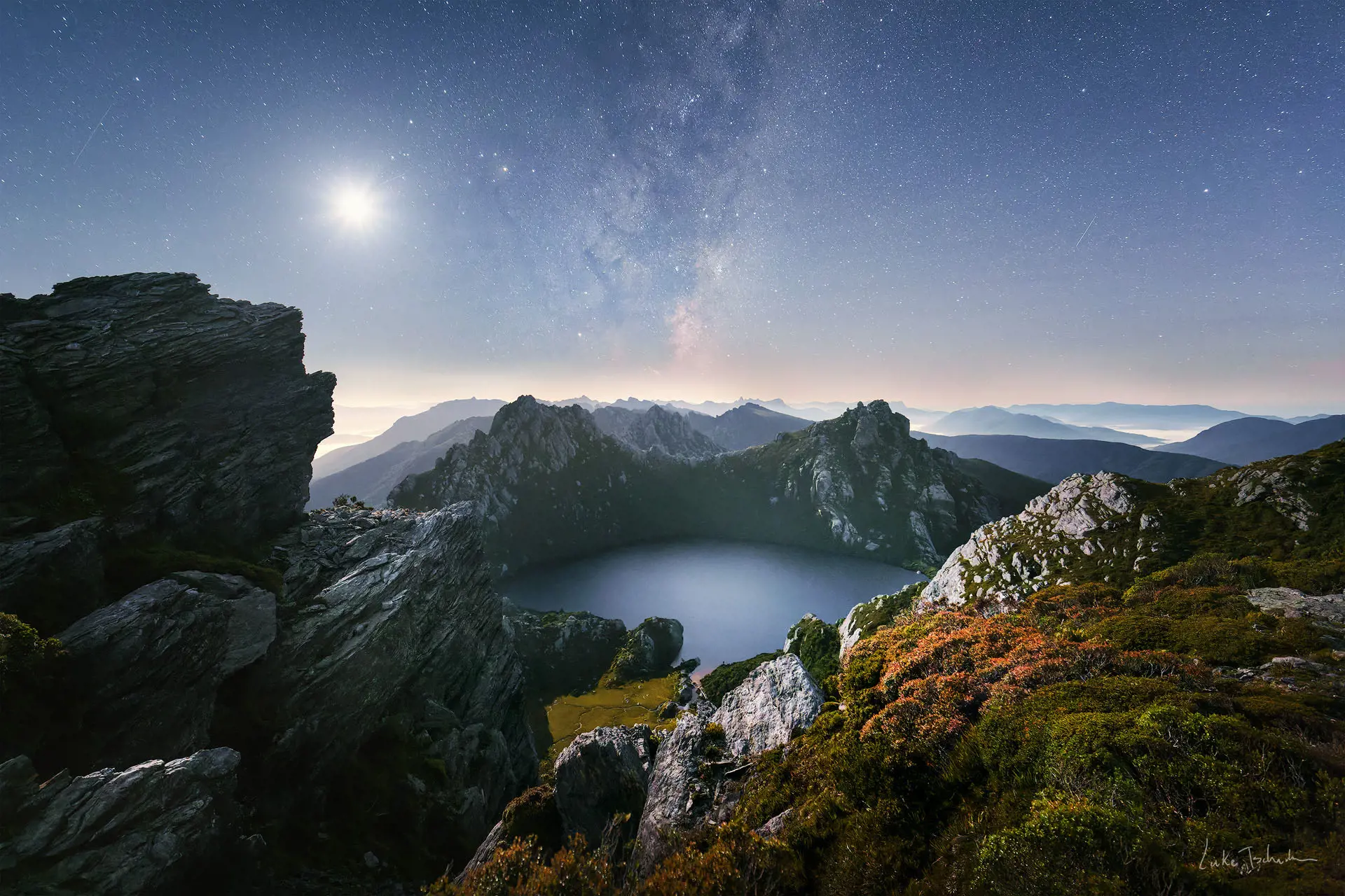A stunning landscape image over the rugged mountains and lakes of south west Tasmania, with the Milky Way and the moon in the brilliant night sky overhead.