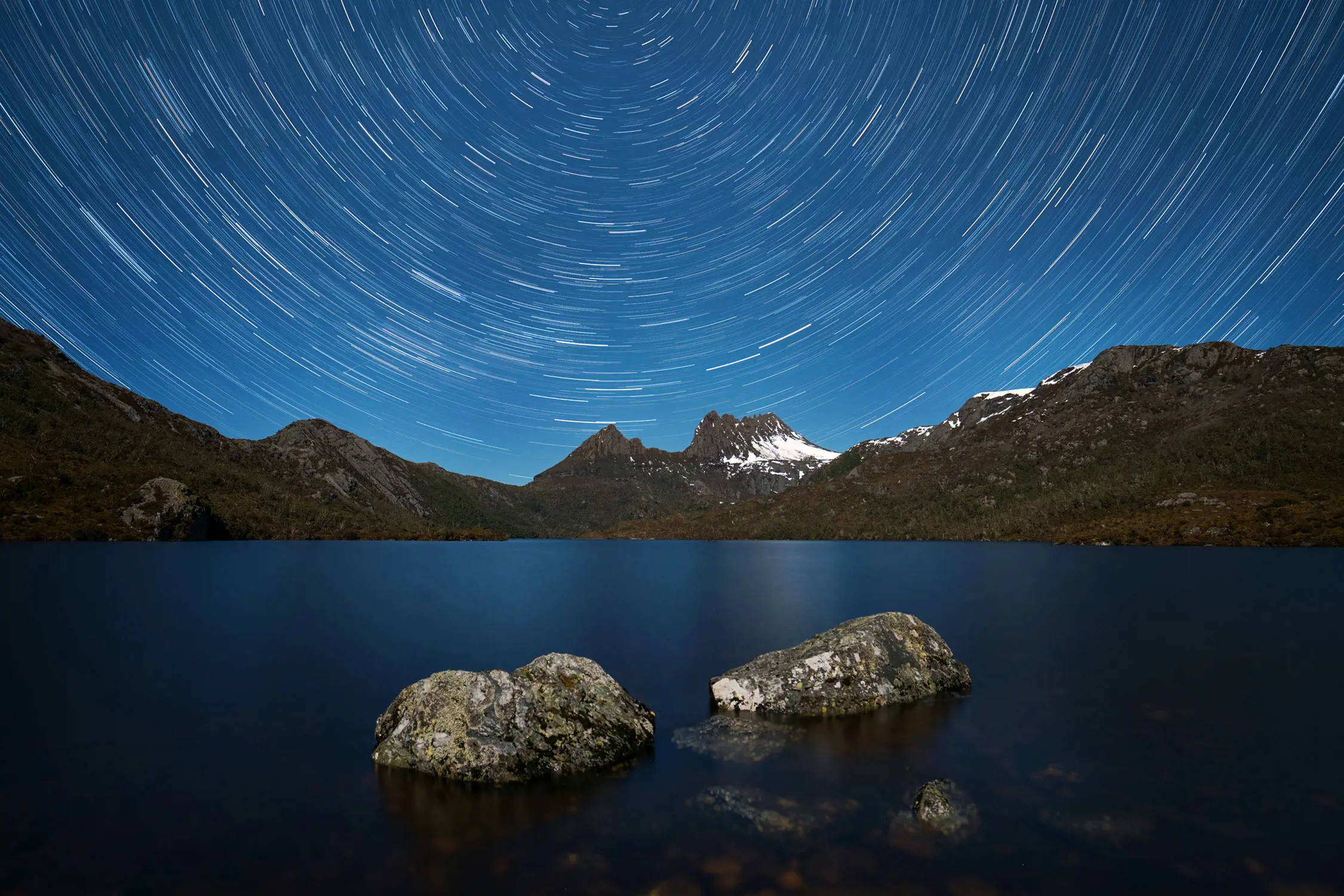 An image of Cradle Mountain and Dove Lake at night. The long exposure of the image creates a swirl of stars in the deep blue night sky