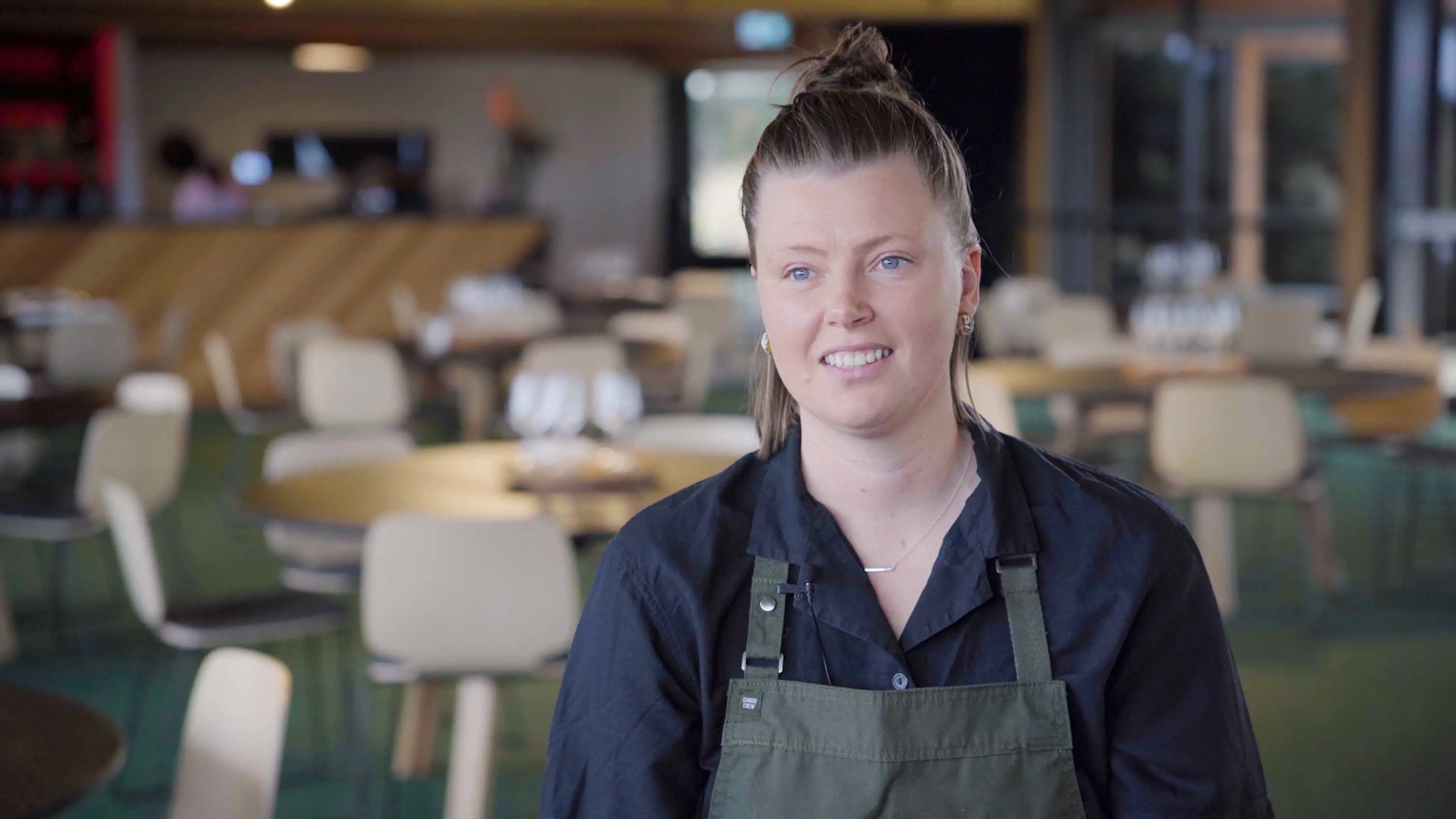 A young woman wearing a blue collared shirt and khaki coloured cooking apron.