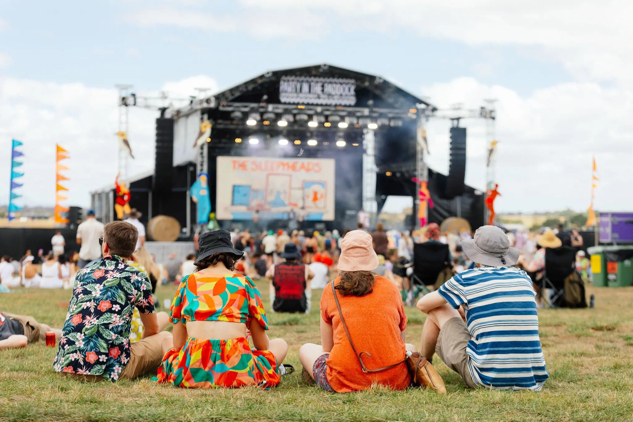Four people in colourful clothes and hats sit on the grass, facing towards a large festival stage with lighting, sound equipment and a banner with 'Party in the Paddock' at the top.
