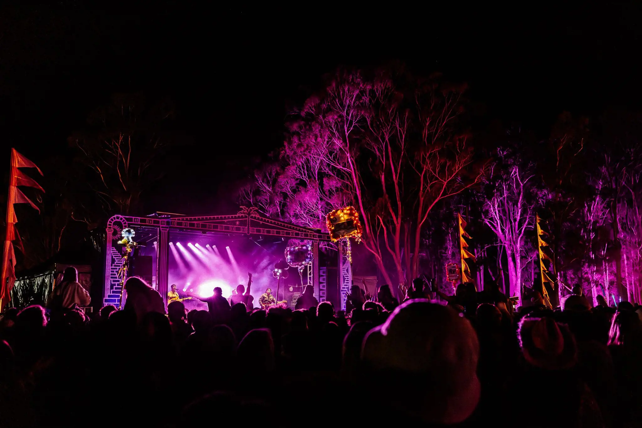A music festival at night. The crowd is illuminated purple from the stage lights, and tall trees reflect the red and purple light.