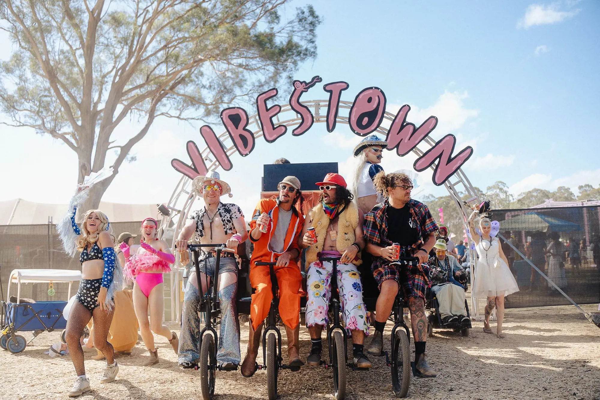 A parade of colourfully and creatively dressed festival attendees. Four people at the front are riding bicycles stacked side by side, and a large archway with 'Vibestown' in pink lettering rises above them.
