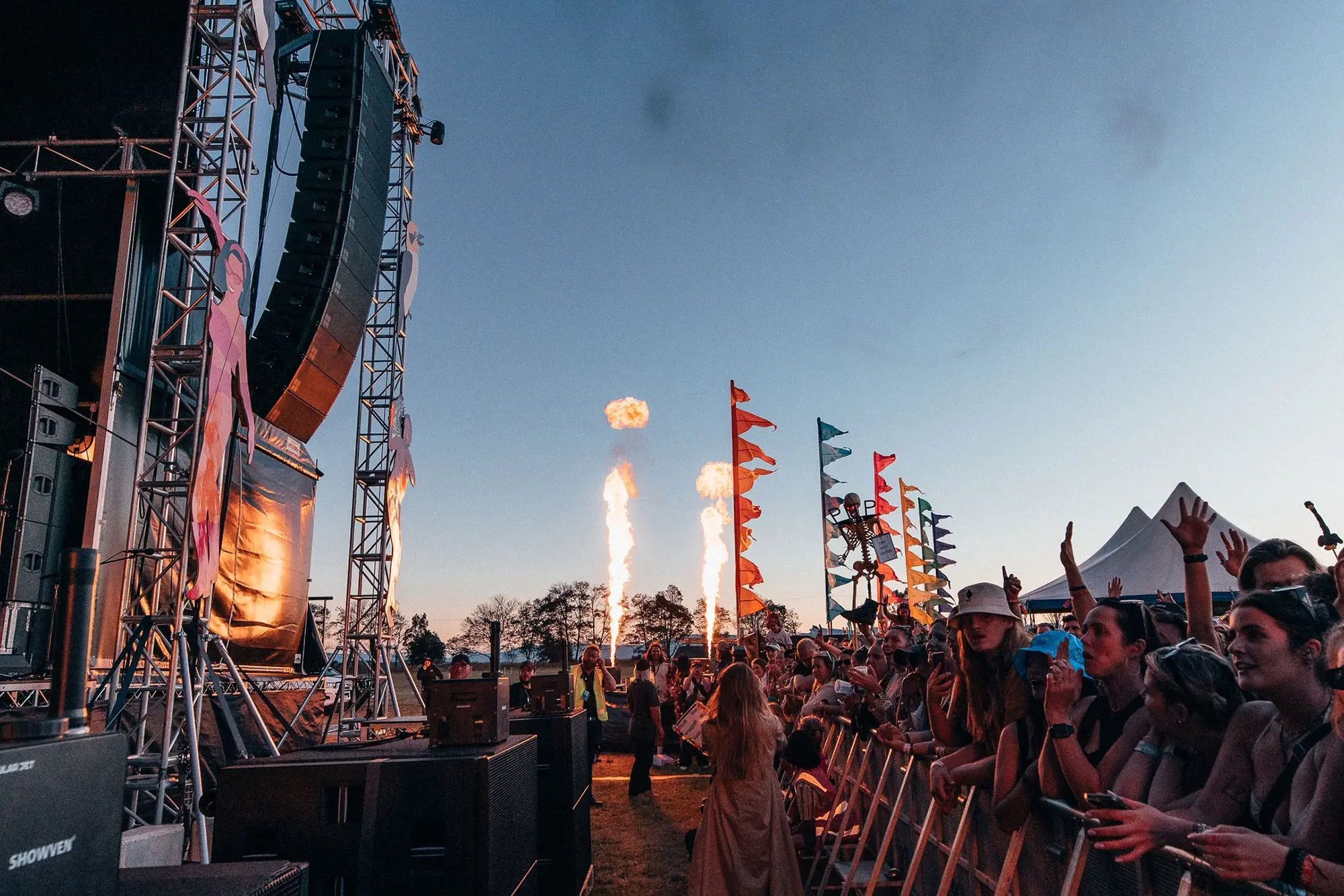 The side-of-stage view of a large festival stage at dusk. The audience lines the barrier, their hands in the air, and colourful banners and pyrotechnics decorate the background.