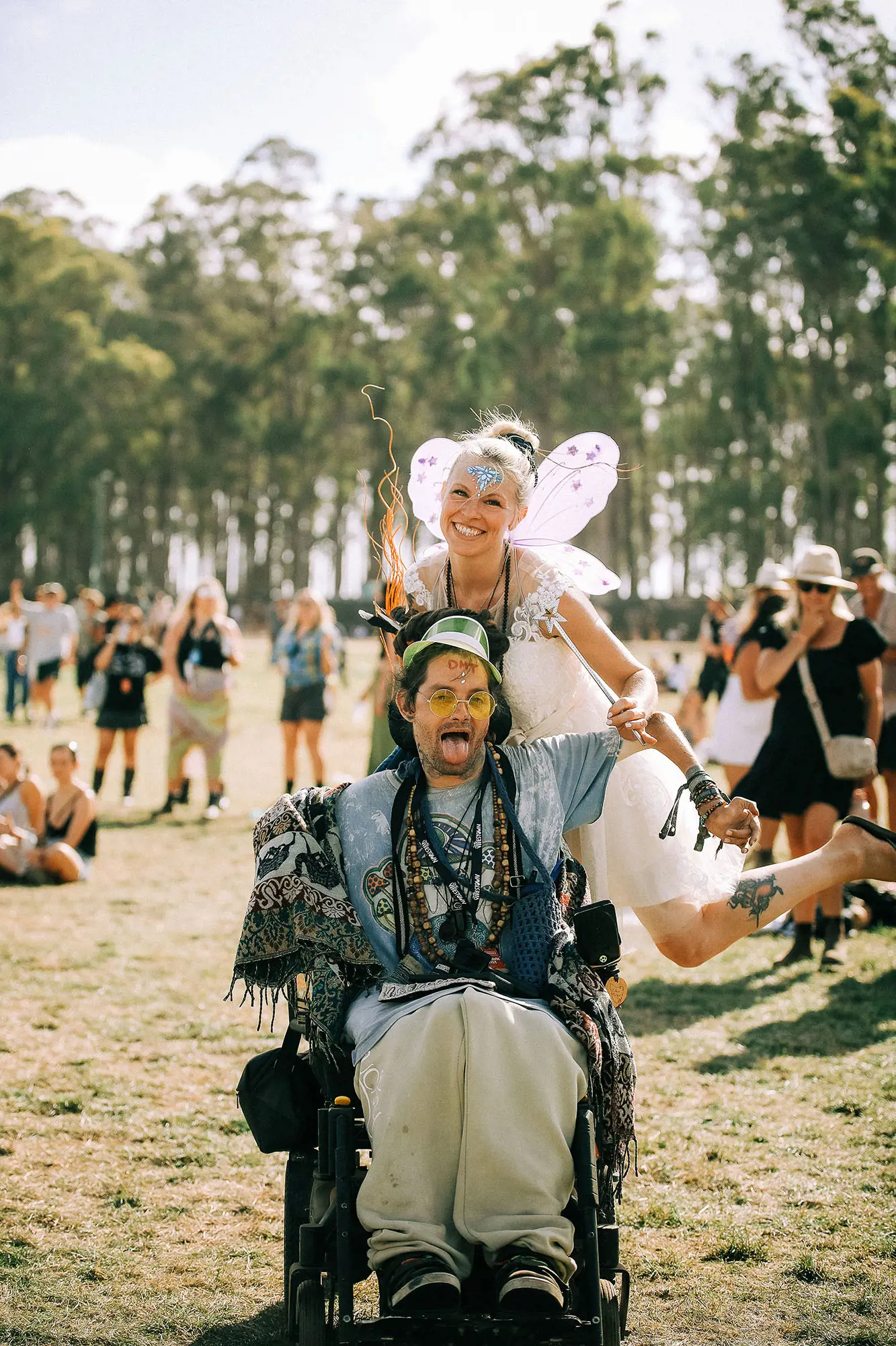 Two people in the middle of a paddock, posing cheekily for the camera. One is in an electric wheelchair, draped in blankets, beads, bracelets and wearing yellow round glasses. A woman stands behind him, wearing fairy wings and a white dress holding a wand.