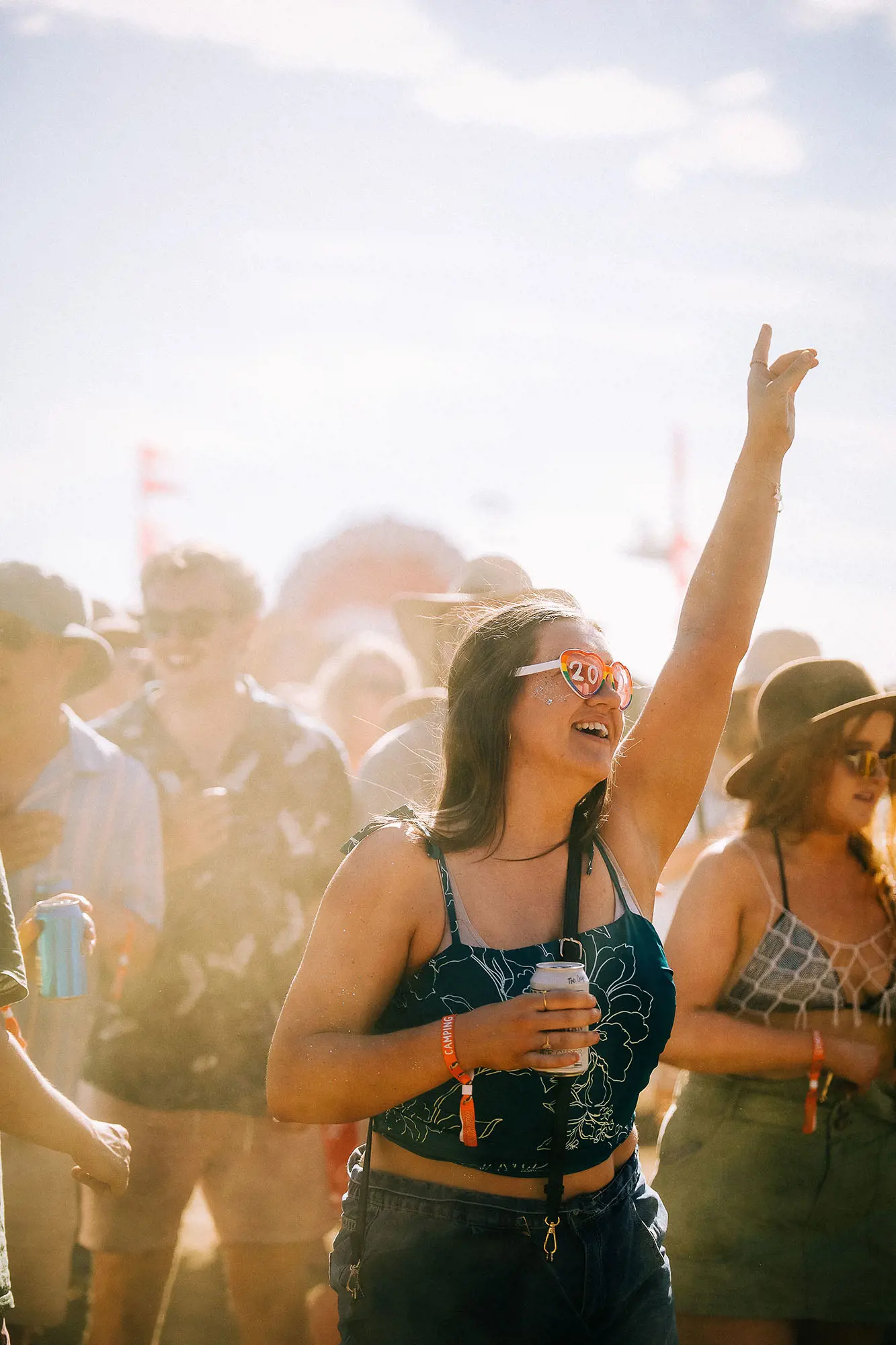 A festival attendee stands amidst a crowd of people, grinning with her hand up mid-dance. She has heart-shaped glasses with the date '2025' across the lenses.