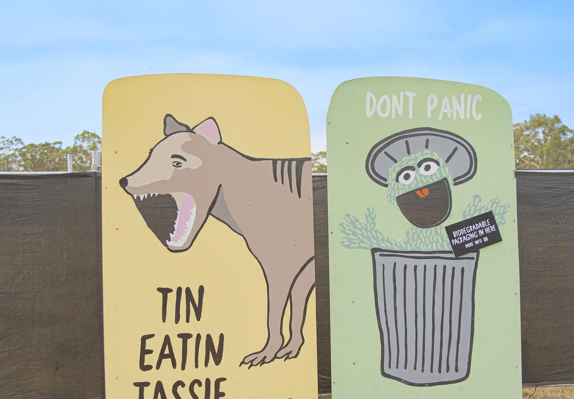 Two signs propped up against a fence. One is yellow, has a Tasmanian tiger and says 'Tin Eatin Tassie'. The other is green, has Oscar the Grouch in a silver rubbish can and says 'Don't panic - Biodegradable packaging in here'.