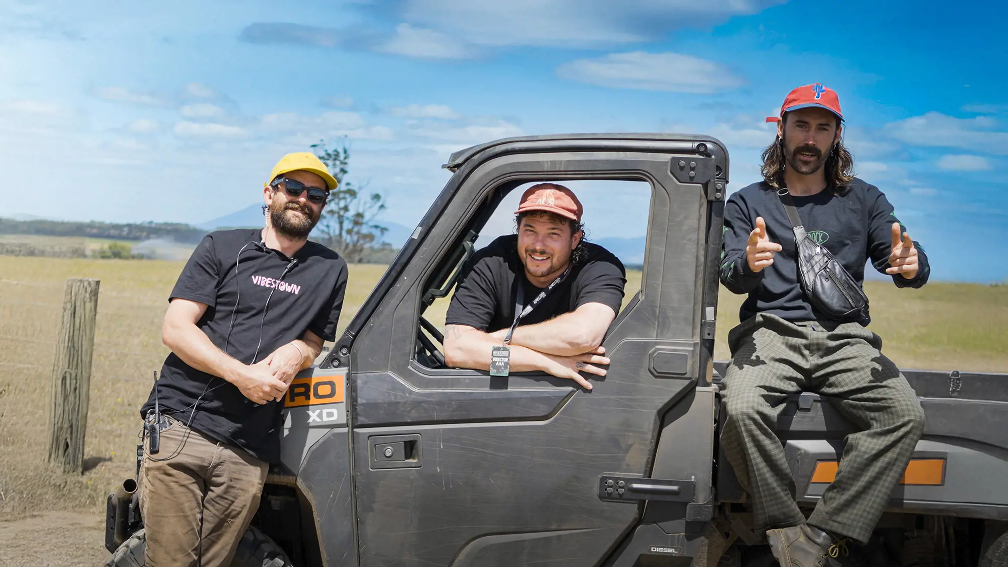 Three men wearing hats, lanyards, radios and black t-shirts pose in and up against a dusty black ATV.