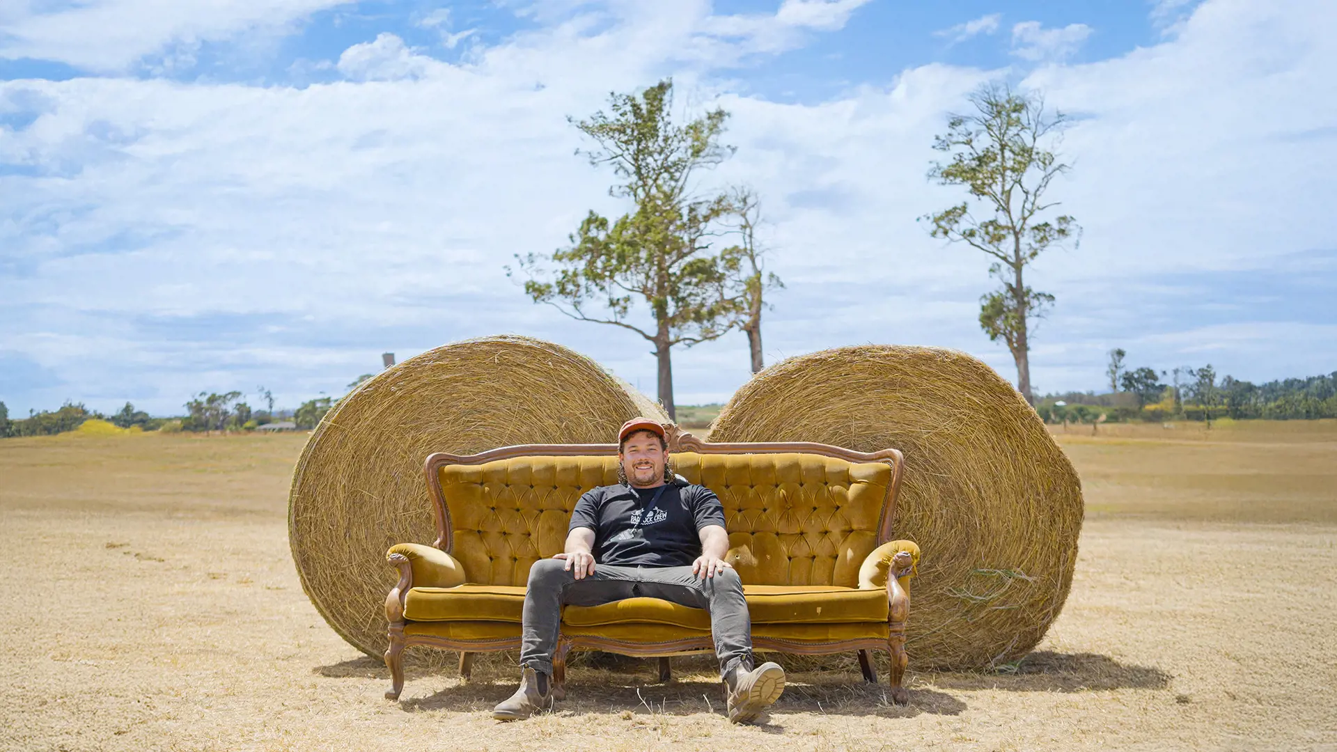 A man in jeans, boots and a black t-shirt sprawls out on a vintage yellow velvet couch, sitting in front of two large hay bales in the middle of an open paddock.