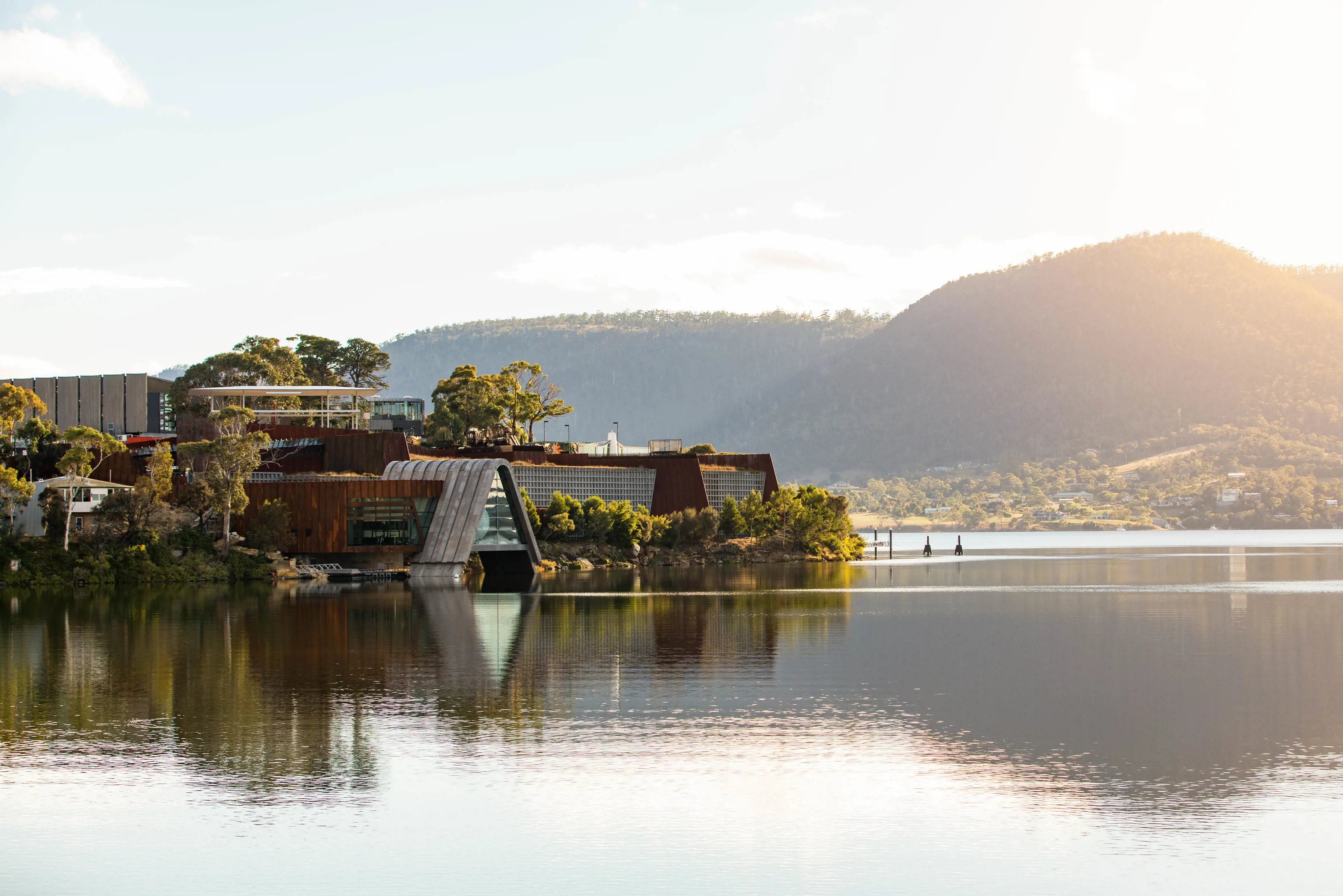 The architectural, unusually-shaped collection of buildings that make up Mona, made from steel, glass and rusted iron. The museum sits amongst trees on land jutting out into a river. The water is calm and still and mountains rise in the background.