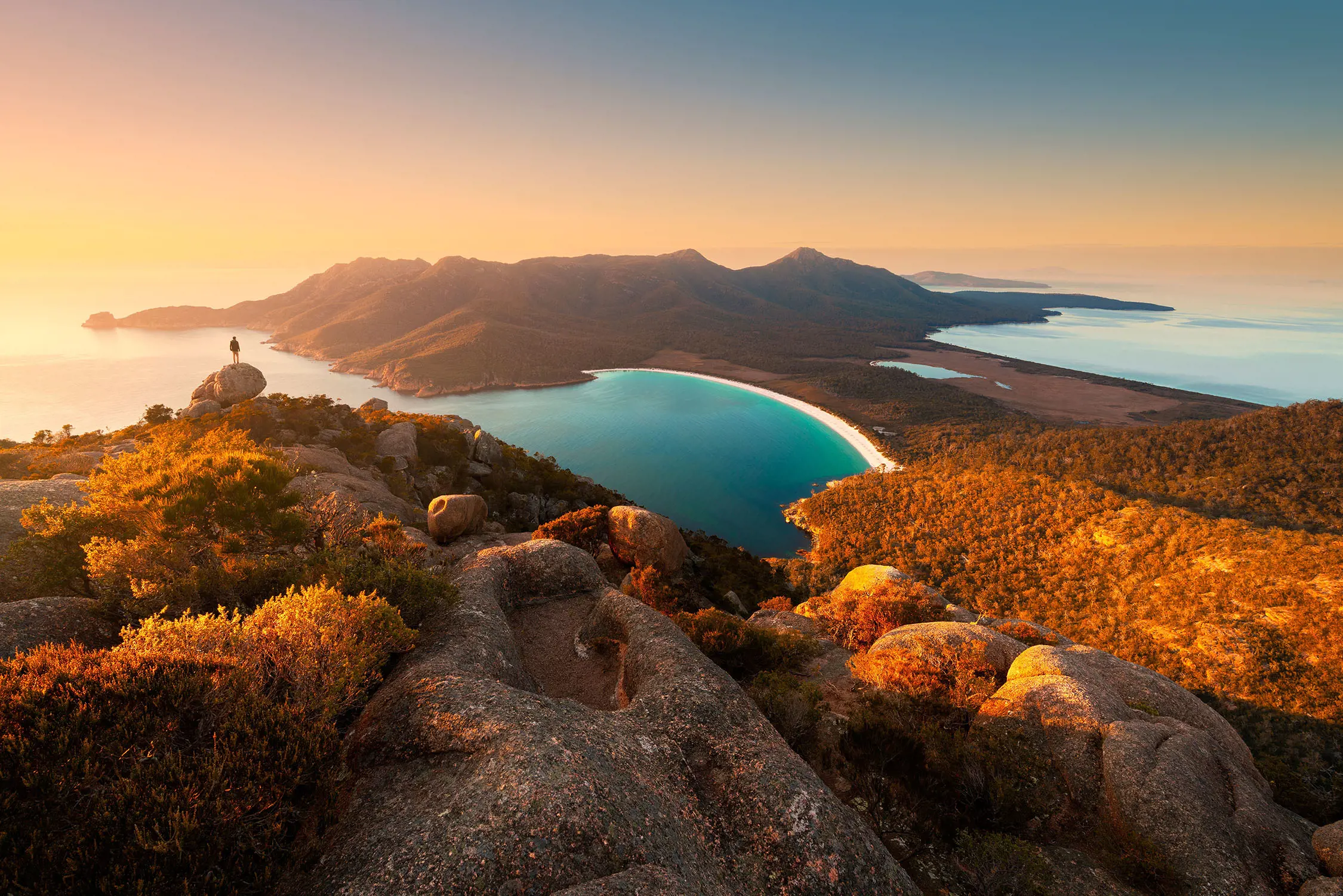 Aerial shot of Wineglass Bay, Freycinet National Park. A man standing on the cliff, looking out onto the water.