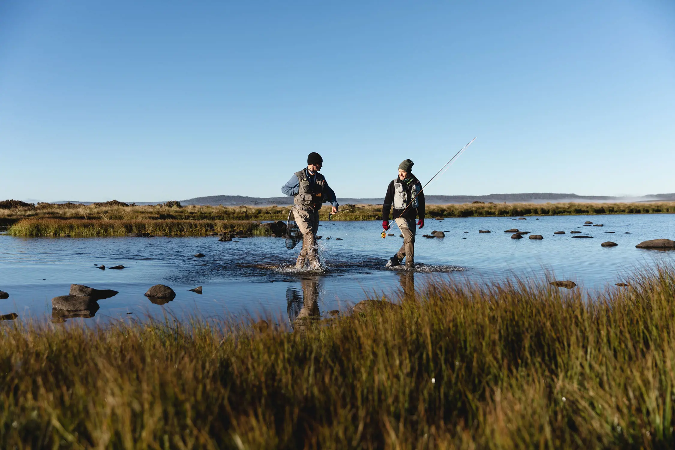 Two people in waders walk through shin-deep lake water, carrying fishing rods and gear. Grassy plains open up around them.