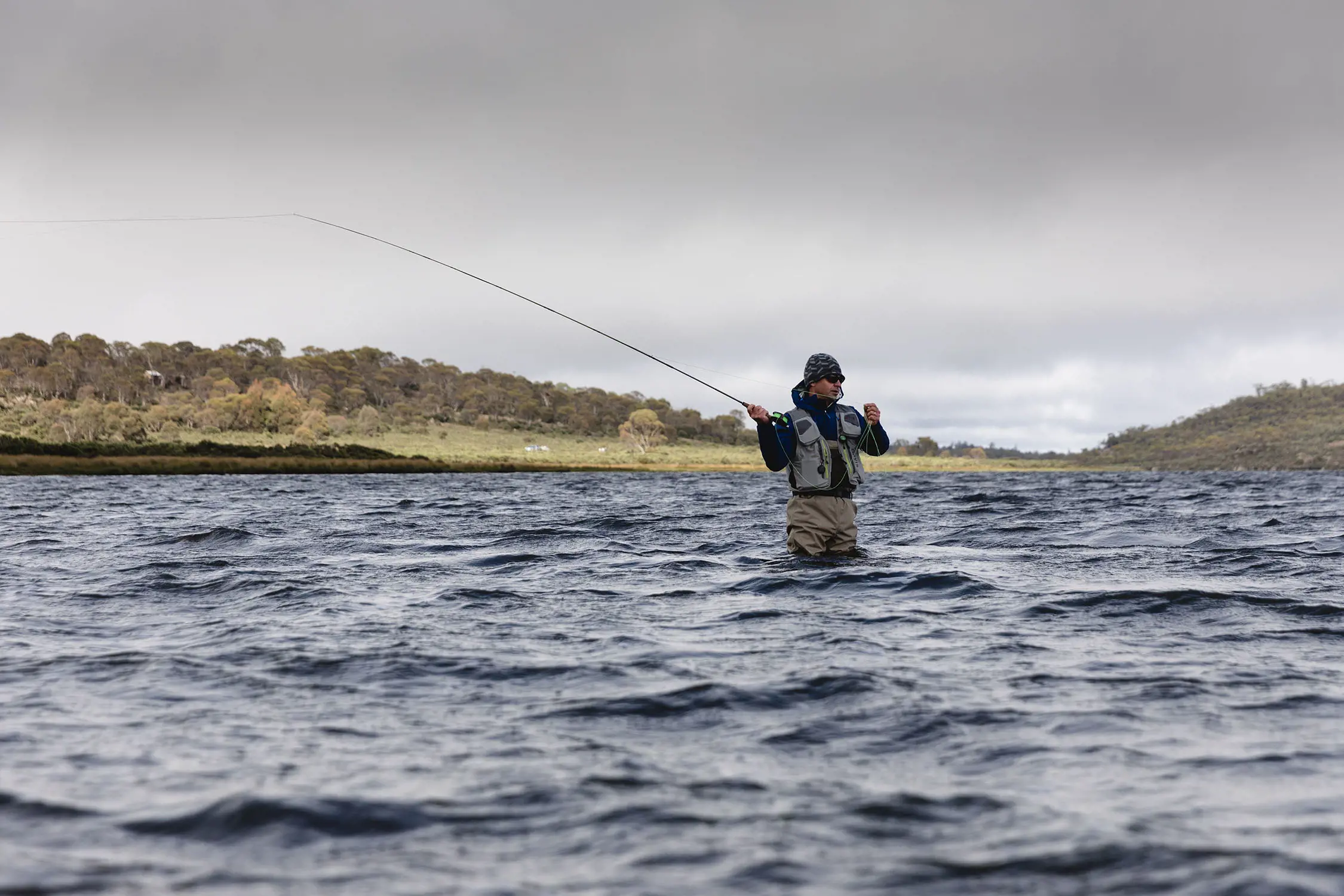 A person in outdoor fishing gear stands in thigh-deep, choppy lake water, with a fishing rod flung back mid-cast.