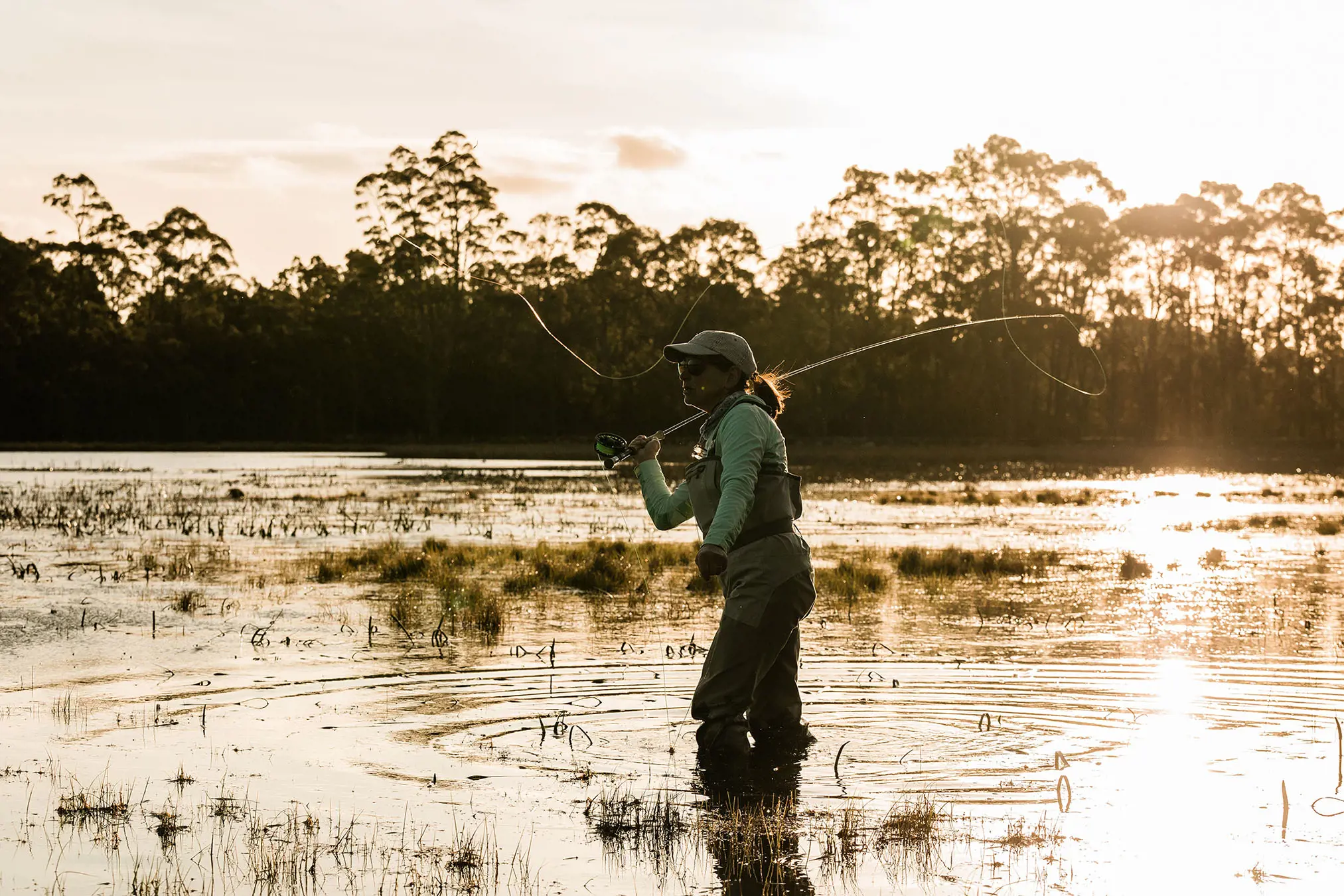 A woman in waders stands in knee-deep lake water, rod and line thrown in the air mid-cast. The sun shines through the bush behind her and reflects off the still water.