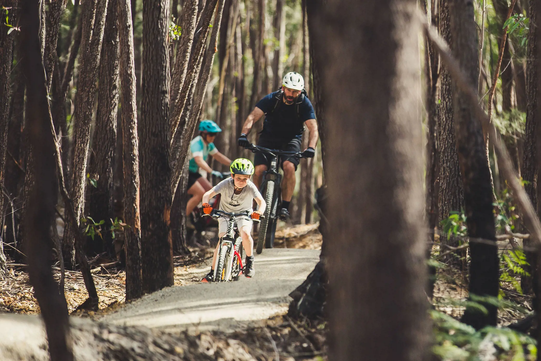 A child and two adults ride mountain bikes along a dirt path winding through trees.