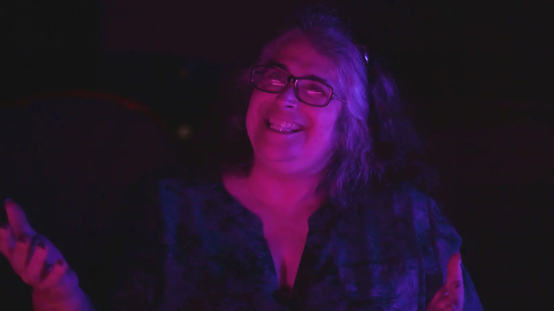A woman with glasses stands in the dark, her face illuminated by a soft pinky-purple glow. She is grinning and mid-gesture.