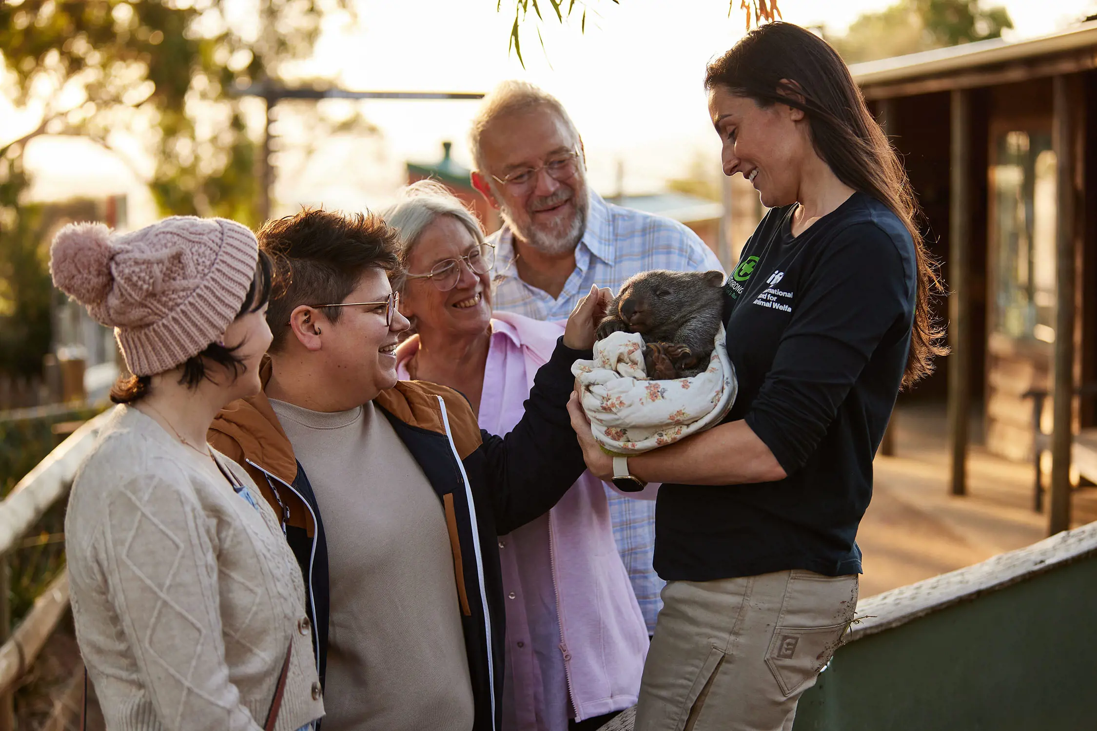 A keeper holds a baby wombat in her arms, snuggled in a patterned pouch, while four people look on eagerly and one reaches out to pat the baby.