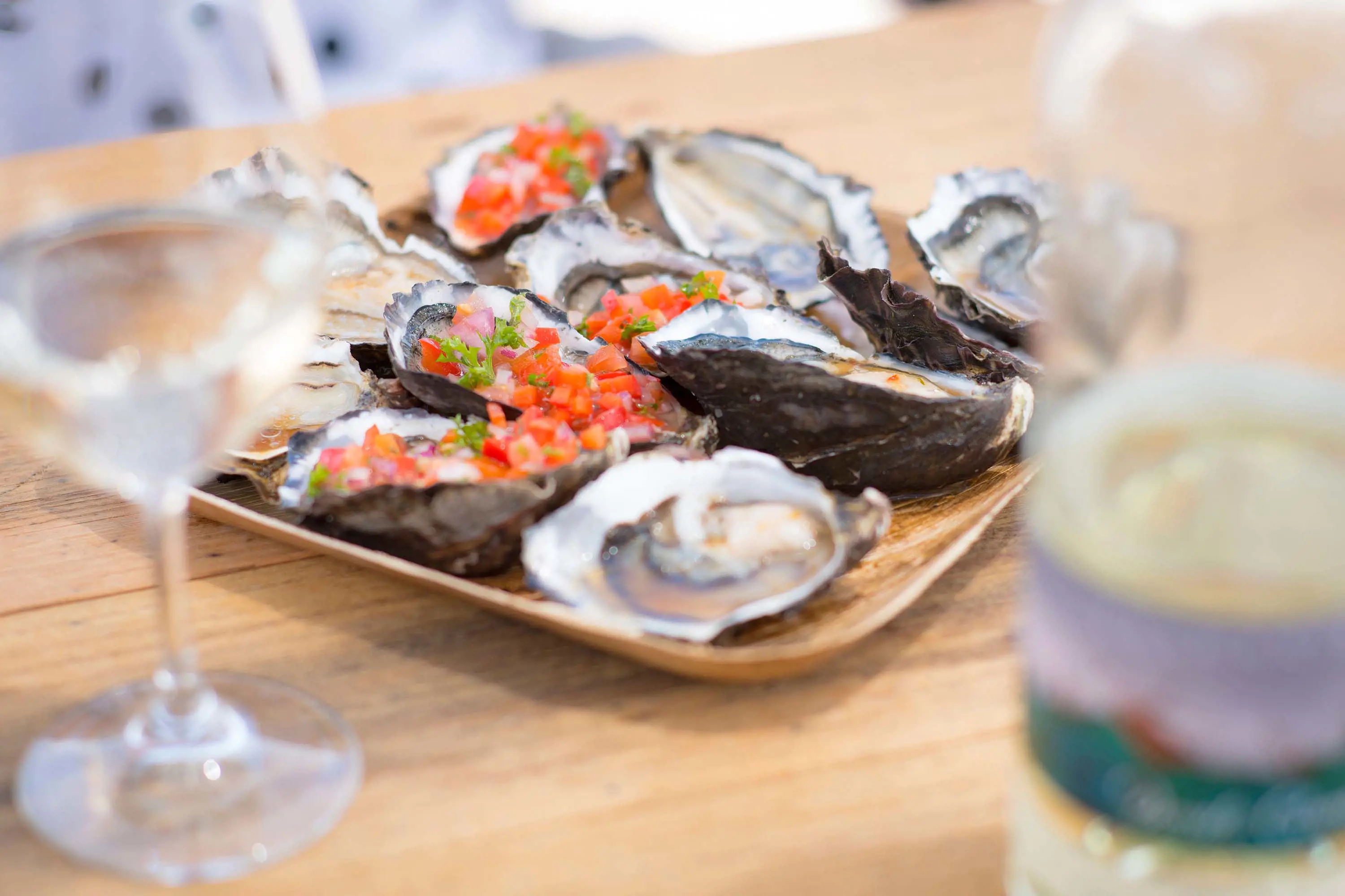 A close-up image of a plate of oysters on a wooden table, with a glass and bottle of white wine.