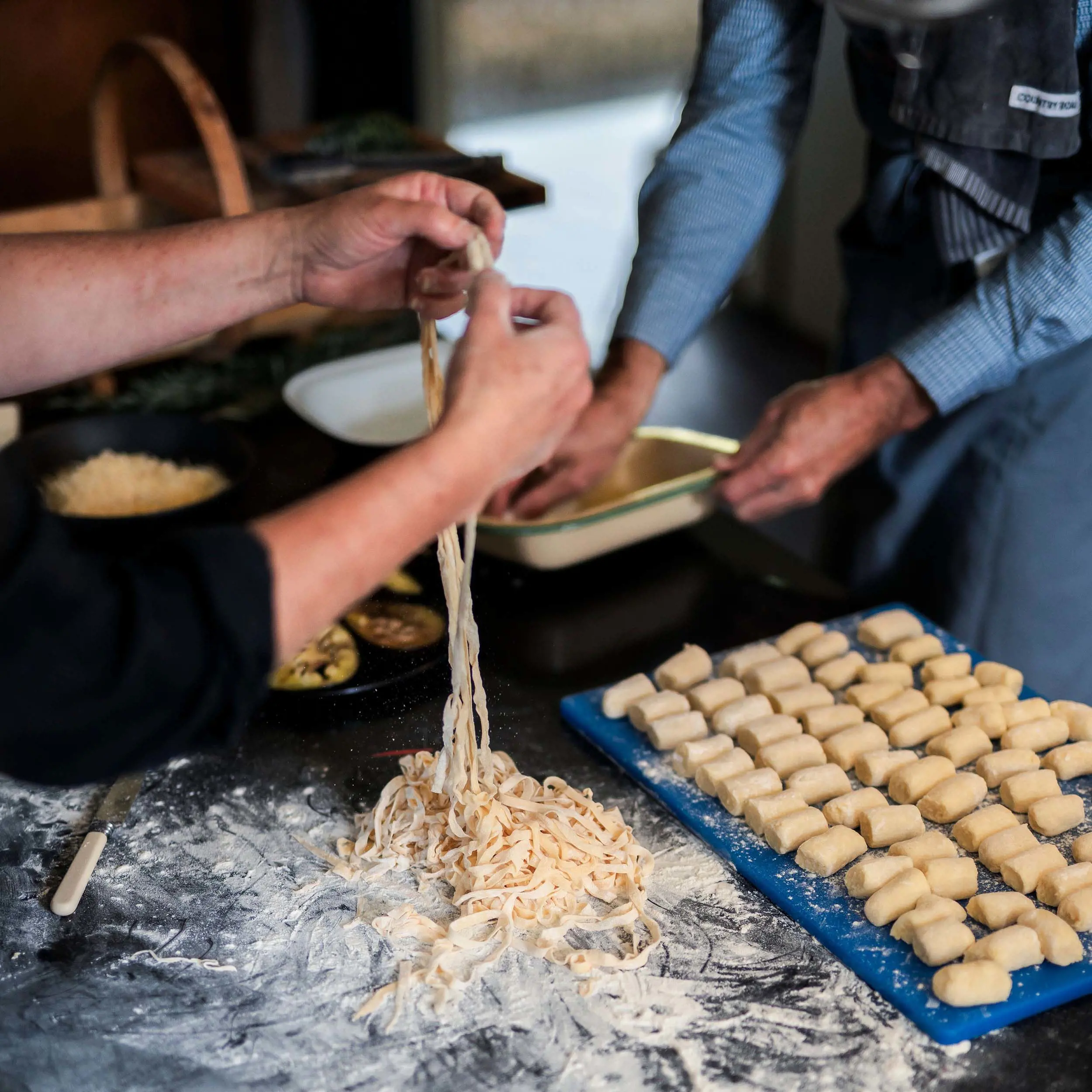 A close-up of hands working in a rustic kitchen. One person stretches fresh handmade pasta, while another shapes small gnocchi pieces, lined up on a floured surface.