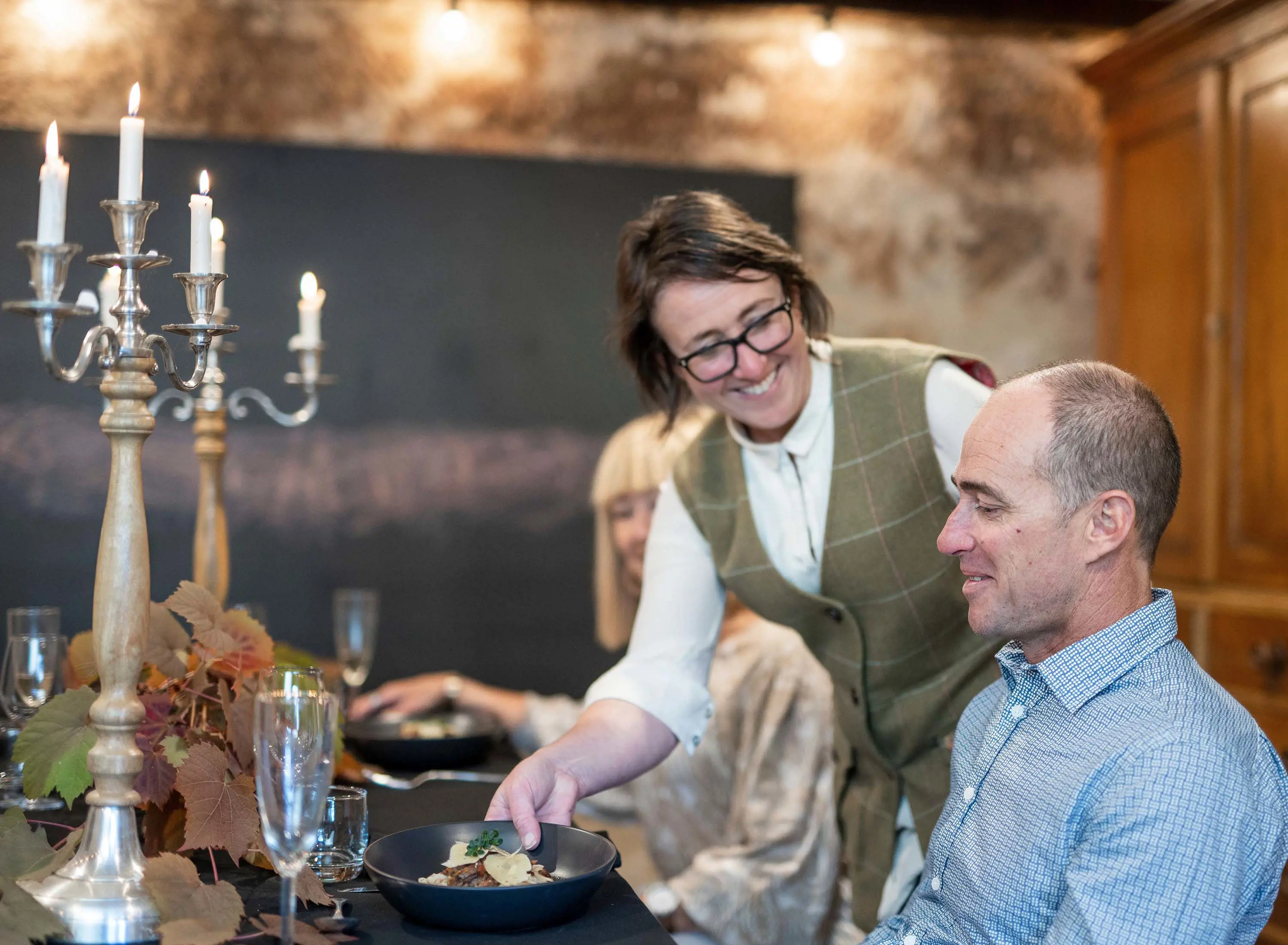 A woman leans over to place a plate of food in front of a man who is seated at a long table, ready to eat. The table is laid with decorations and tall candelabras.