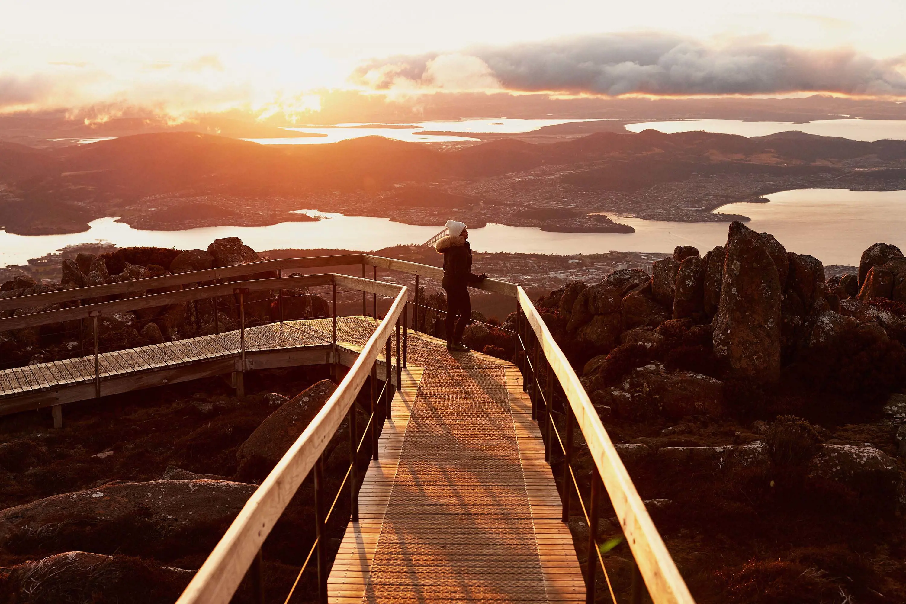 A woman stands at the end of a wooden lookout on top of a mountain, looking down over Hobart.