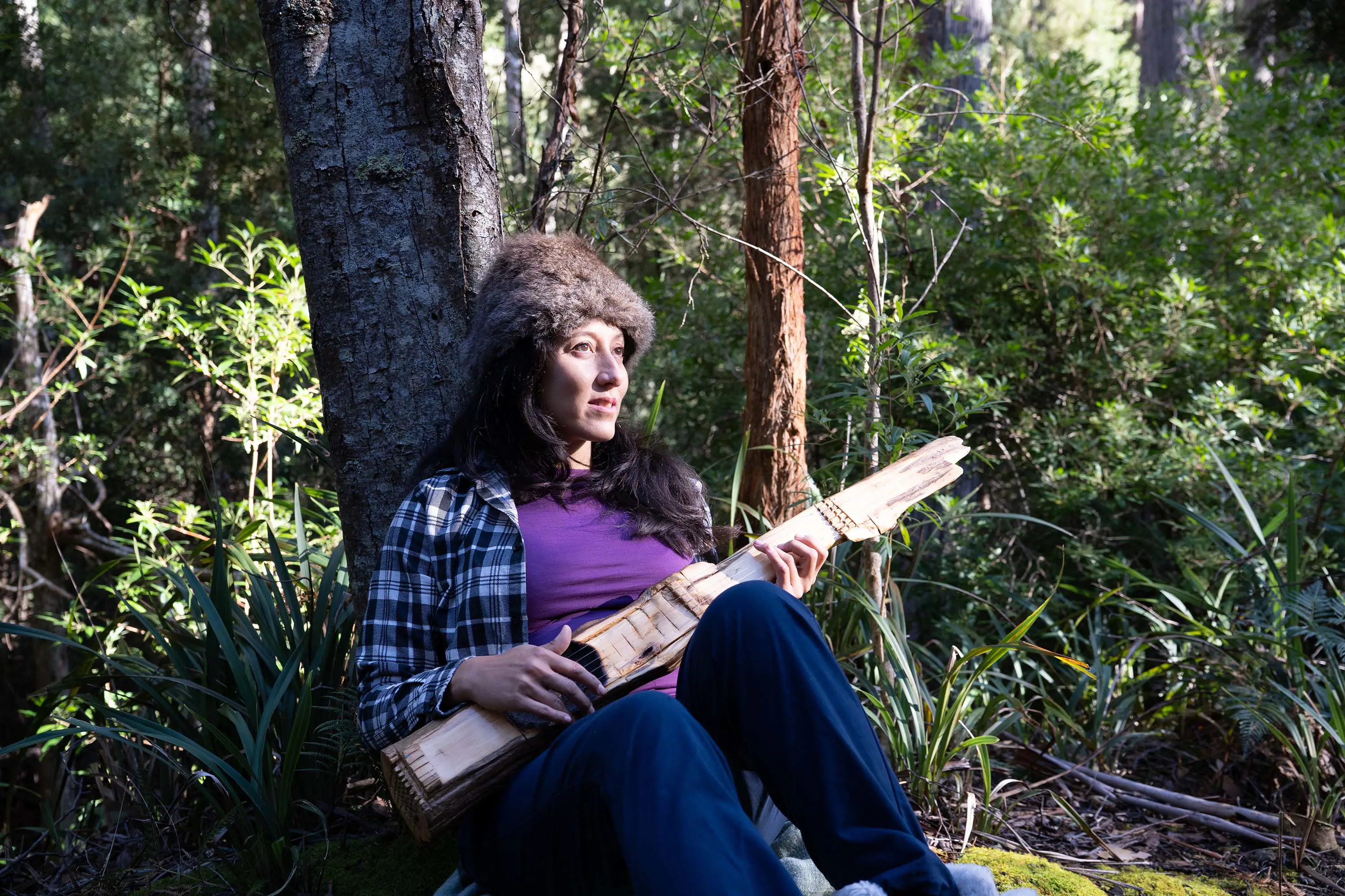 A woman in a furry beanie sits on the ground in the bush, leaning up against a tree. She holds a long piece of driftwood fashioned into a guitar.