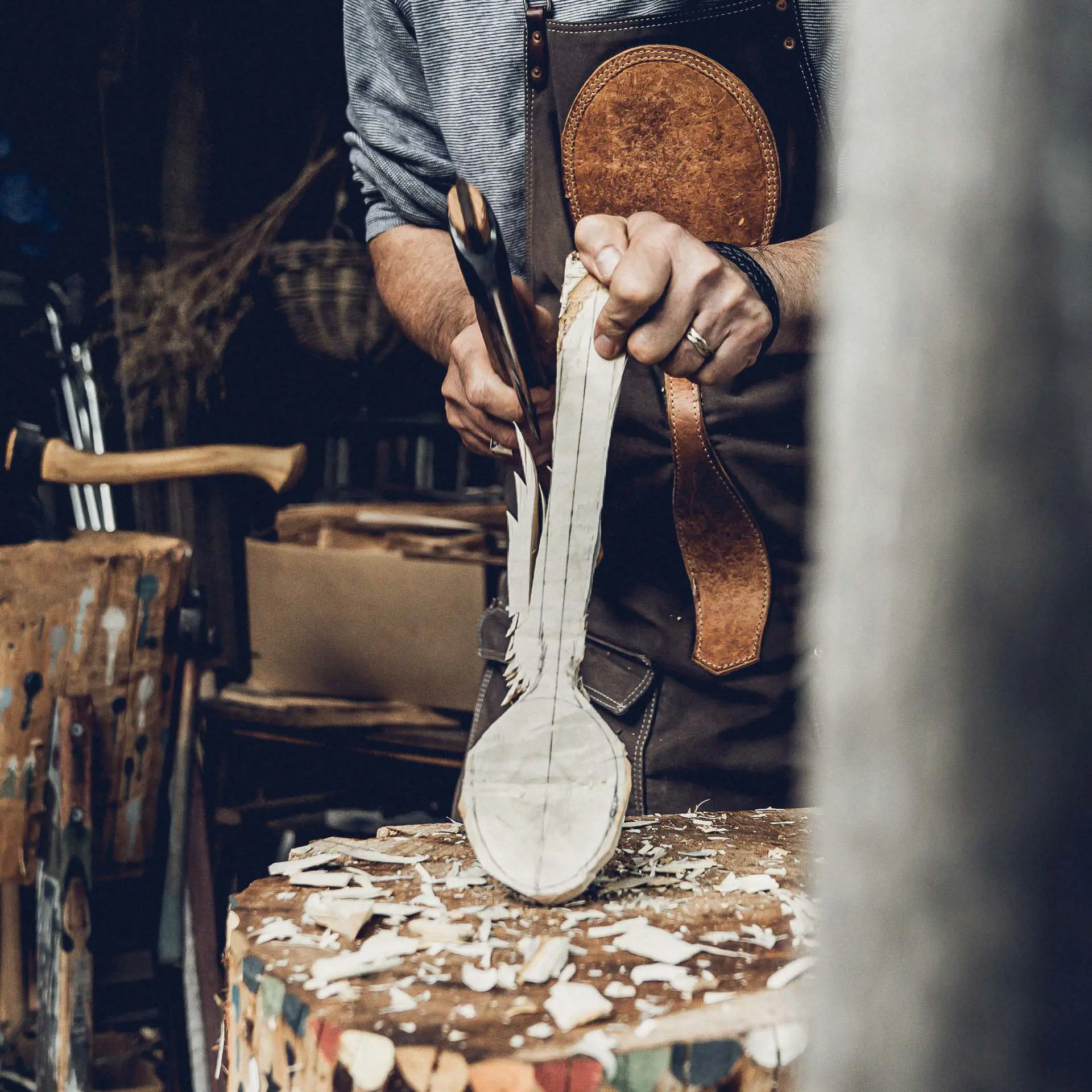In a rustic workshop, a man wearing a leather apron uses an axe to carve the side of a roughly spoon-shaped piece of wood.