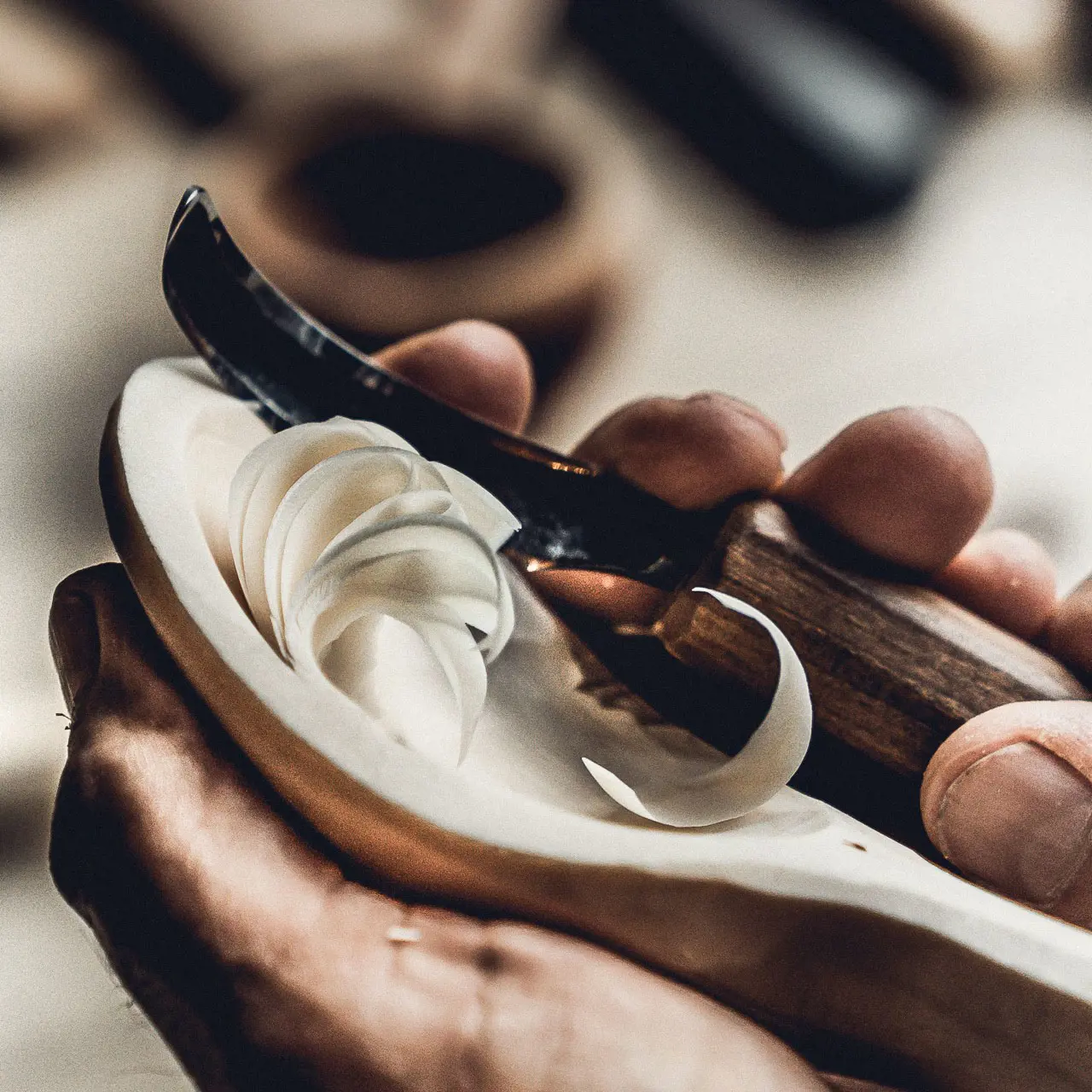 A close-up image of a person's hands holding a piece of wood and a carving knife, carving the wood into a spoon.