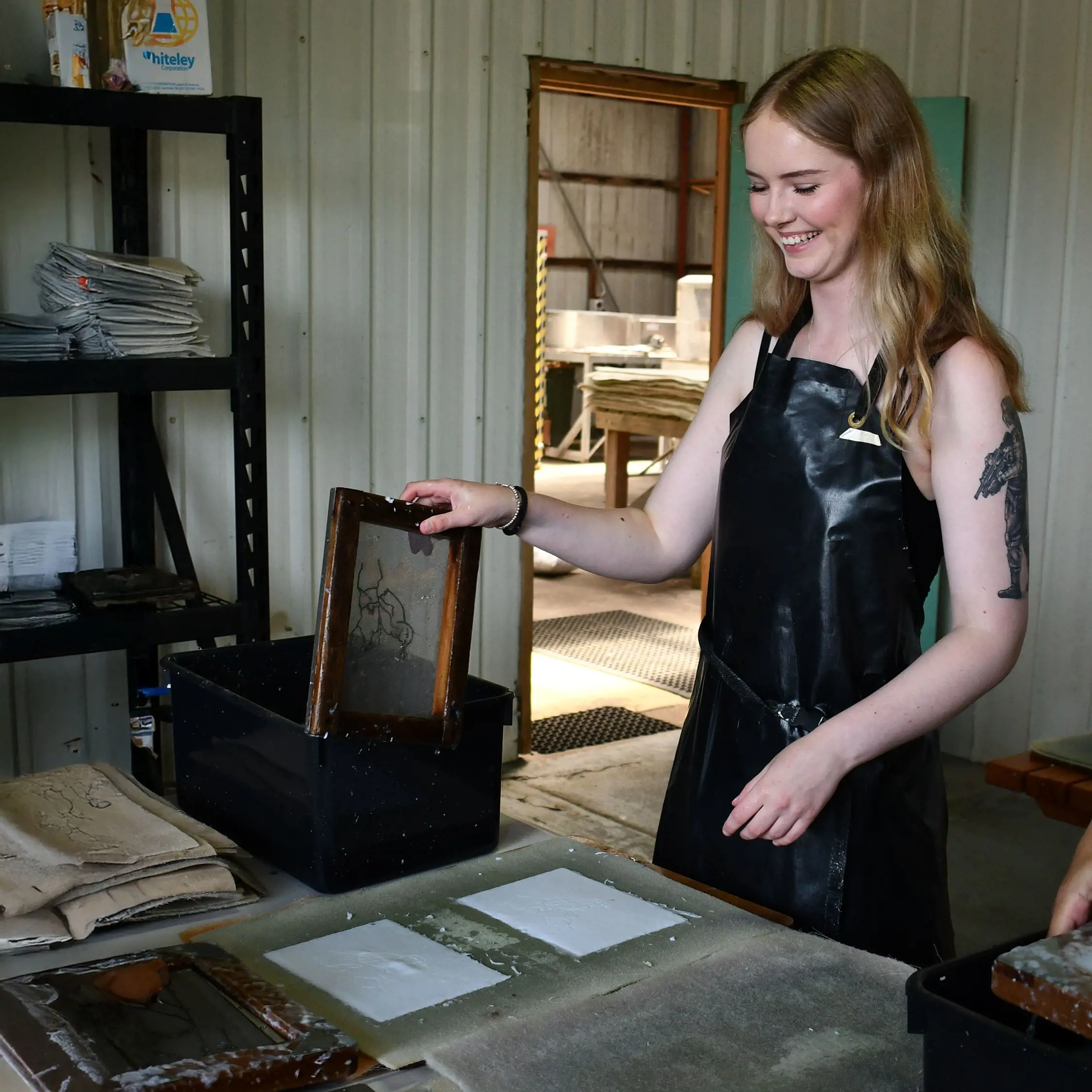 A woman wearing a plastic apron stands in a workshop shed, holding a timber frame used to make paper. Sheets of drying paper are laid out in front of her.