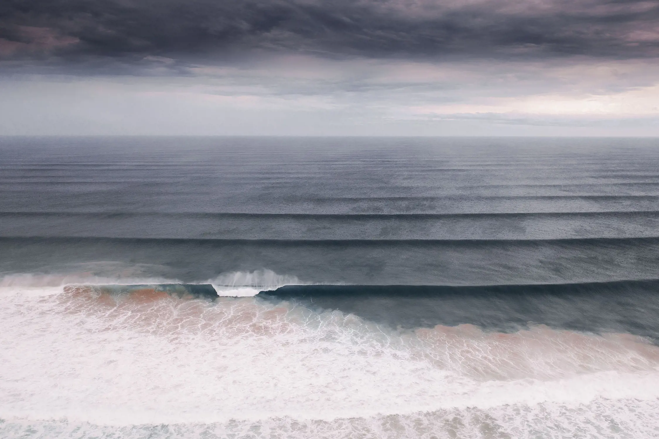 An atmospheric aerial image of a beach. The dark grey water has even waves rolling in and crashing onto the beach, and a heavy grey cloud cover disappears into the horizon.