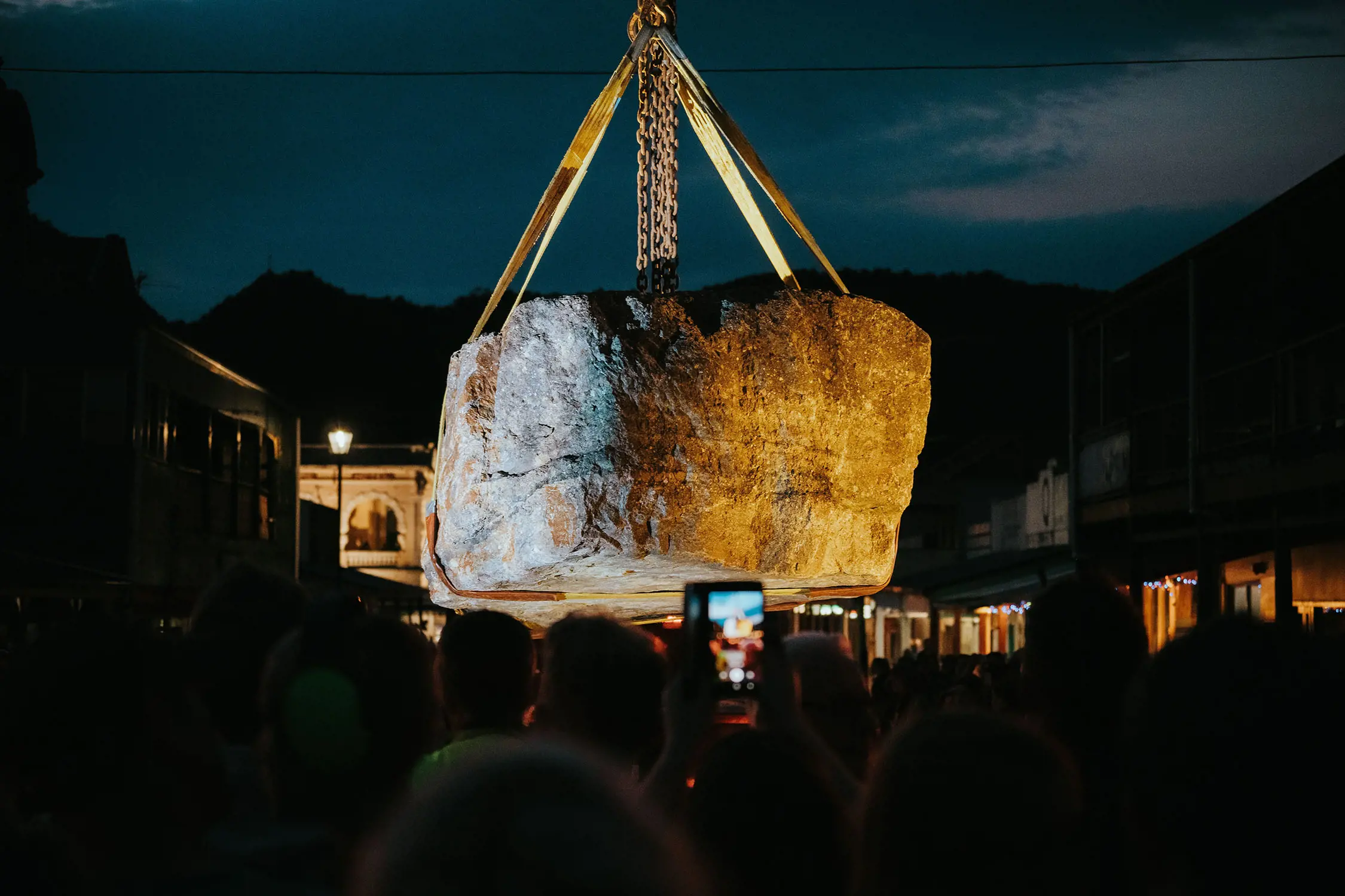 In a crowded town square at night, people watch as a huge illuminated boulder is lifted high into the air.