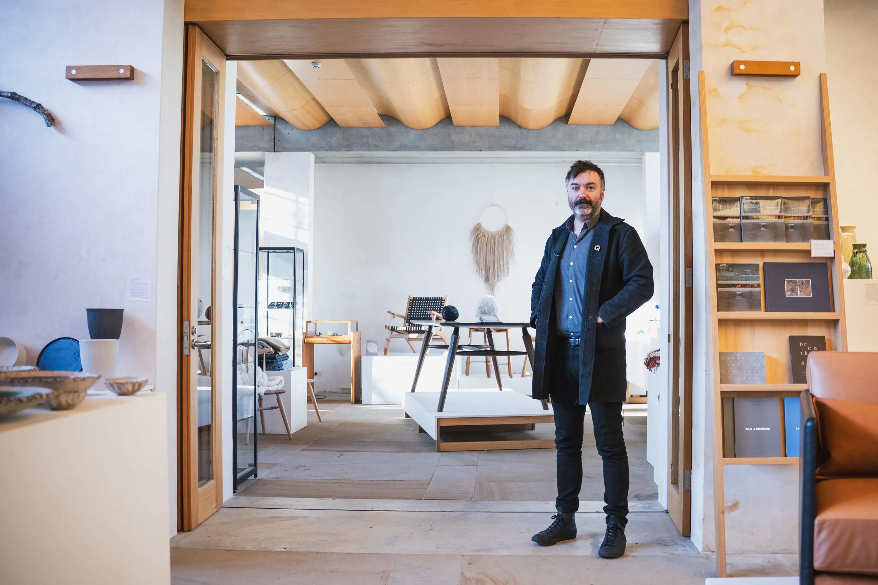 A bearded, middle-aged man in a jacket and black jeans stands in the doorway of a gallery space, looking out onto displays of smooth timber furniture, glass display cabinets and architectural details.