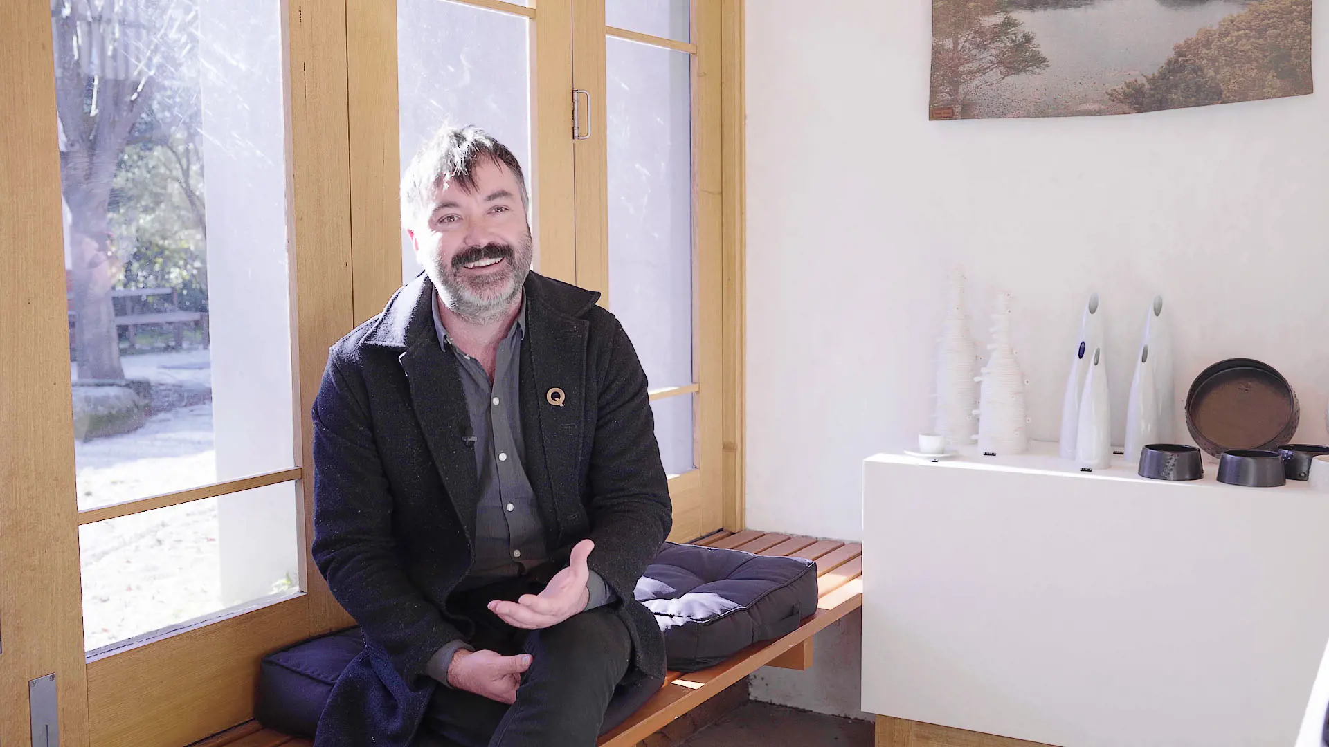 A bearded, middle-aged man in a jacket and black jeans sits on a wooden side bench of a gallery space. Sun streams through the windows behind him, and to his left is a display of sculptural art pieces.